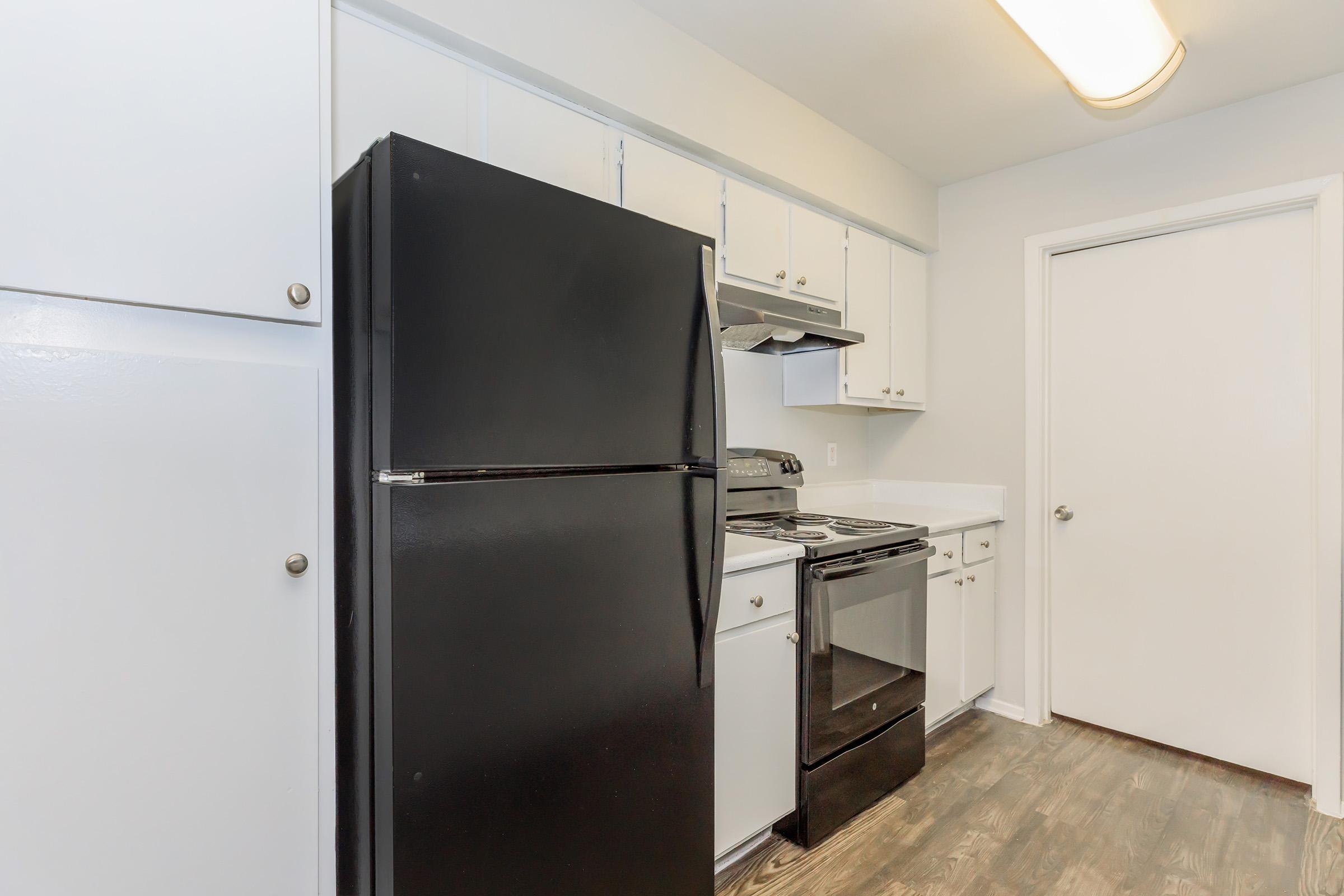 A modern kitchen featuring white cabinets, a black refrigerator, and a black oven range. The layout is clean and spacious, with a door leading to another room. The flooring is wooden, enhancing the contemporary look of the space.