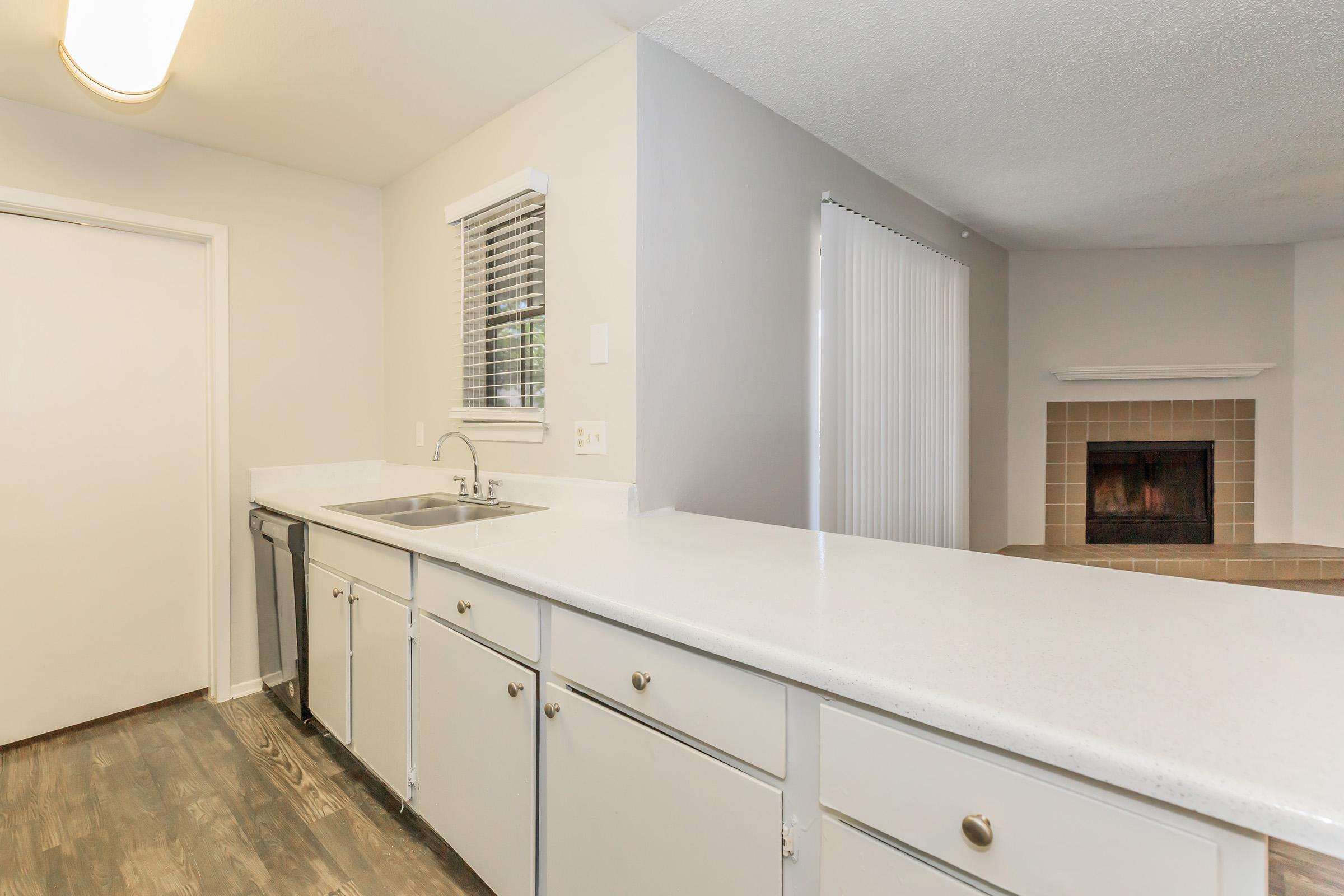 A modern kitchen featuring a white countertop, stainless steel sink, and light cabinetry. In the background, there's a living area with a fireplace and sliding glass doors. Natural light flows in through a window, creating a bright and inviting atmosphere.