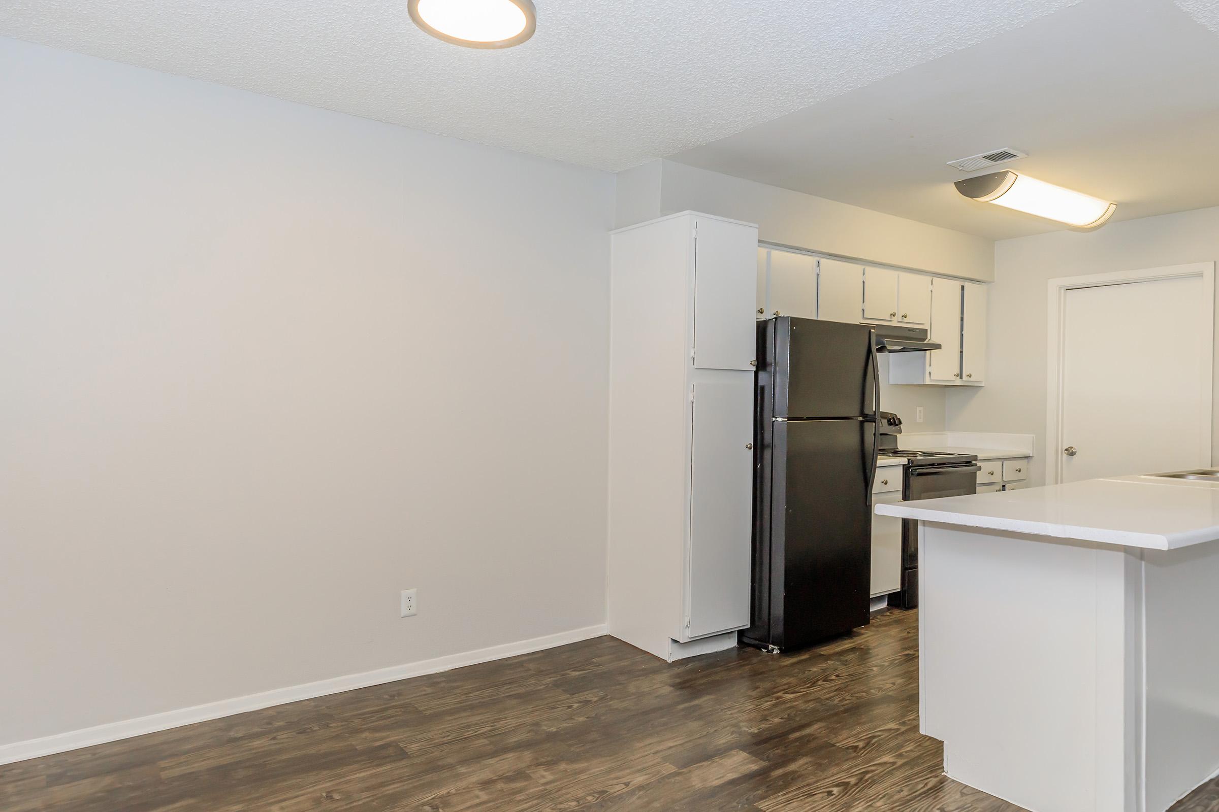 Modern kitchen featuring white cabinets and a black refrigerator. The room has light-colored walls, a light fixture on the ceiling, and dark wood-style flooring. An island with a white countertop is visible, along with a stove and an opening leading to another space.