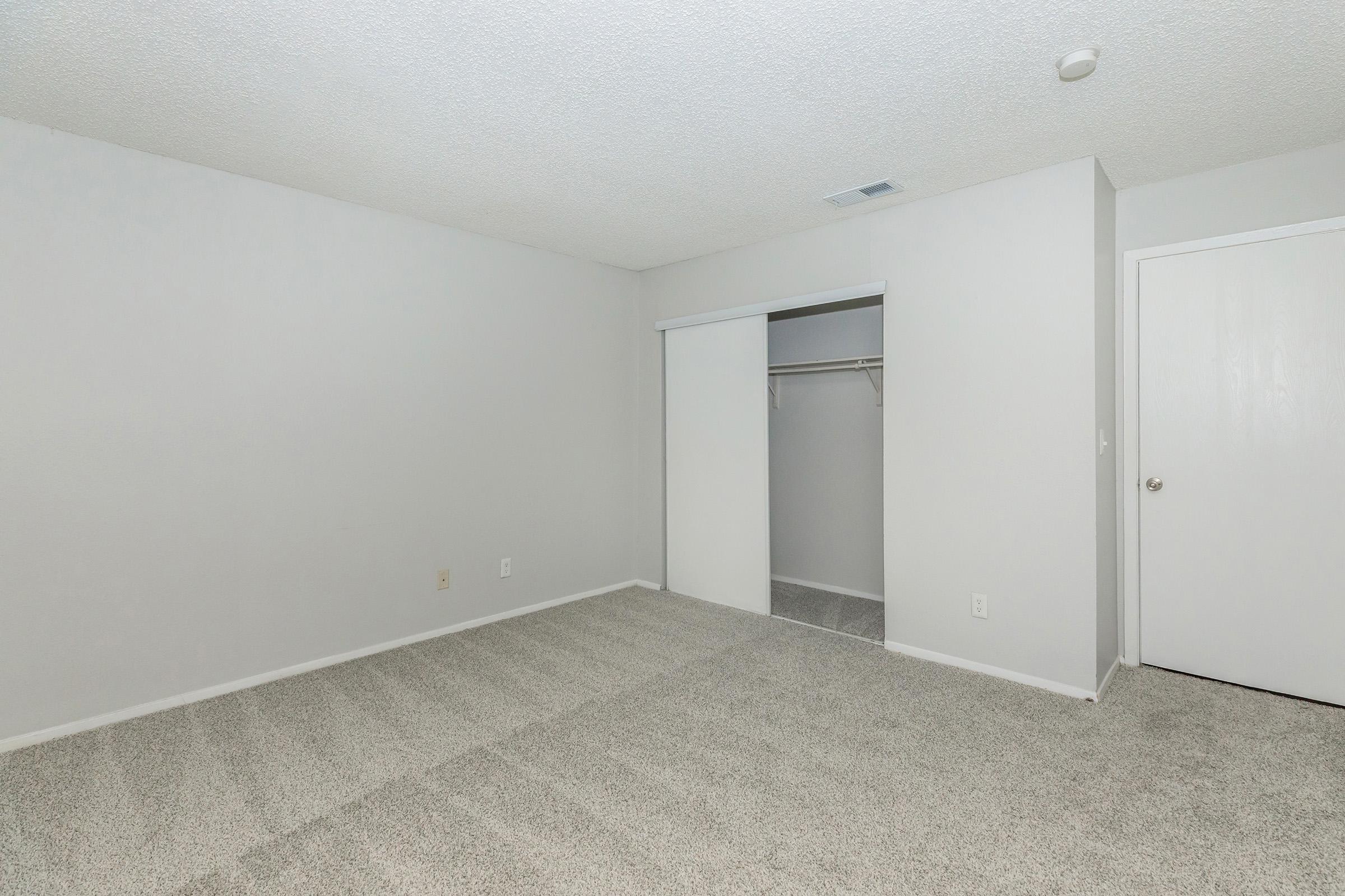 Empty room with light gray walls and a textured ceiling. The floor is covered with short, light-colored carpet. There is a closet with sliding doors on one wall and a white door leading to another area. The space is bright and spacious, devoid of furniture or decorations.