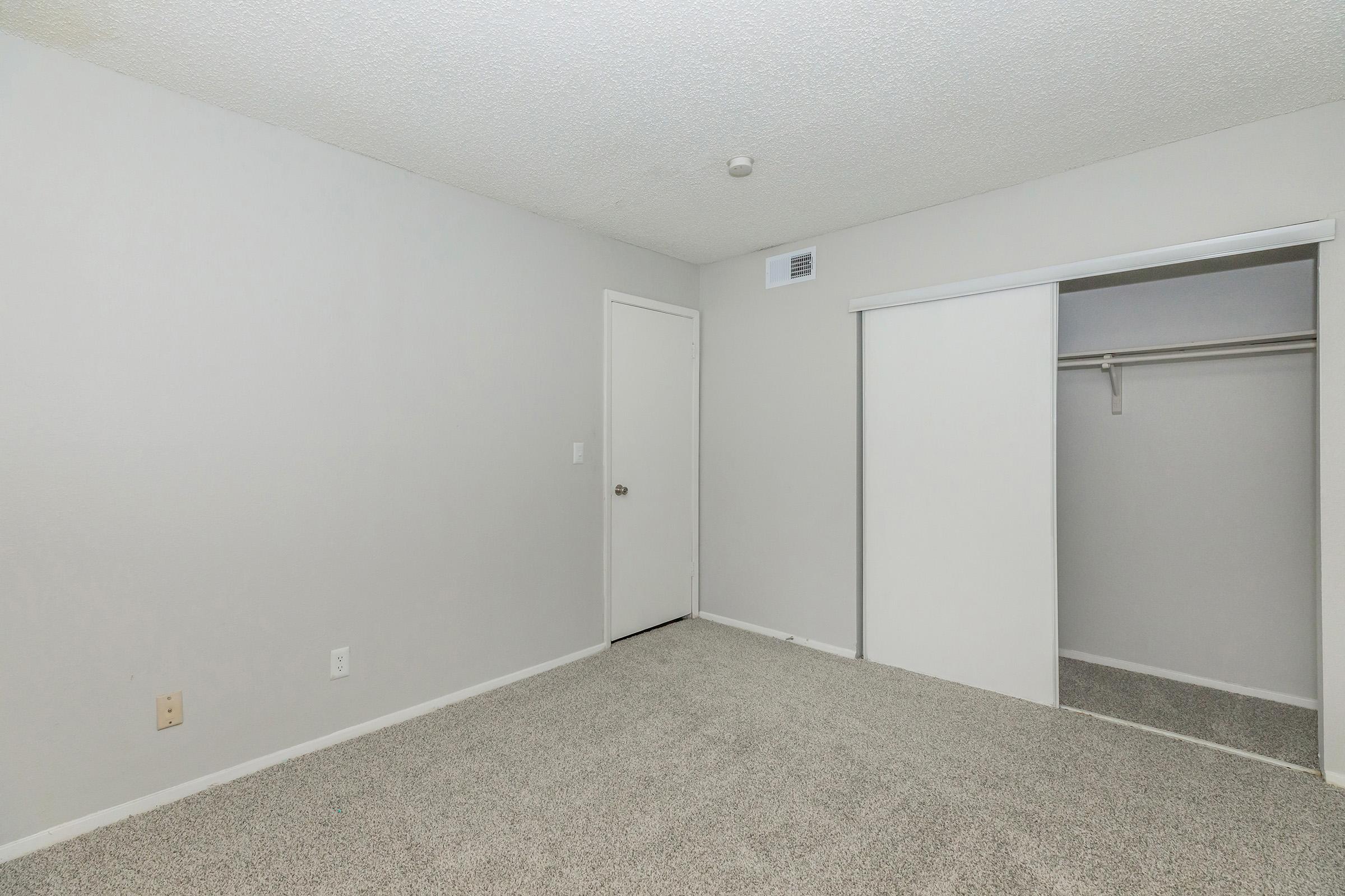 Empty room with light gray walls and carpet. A closed door is visible on the left side, and a closet with sliding doors is on the right. The room is well-lit, and there are no furnishings or decorations, providing a clean and minimalistic appearance.