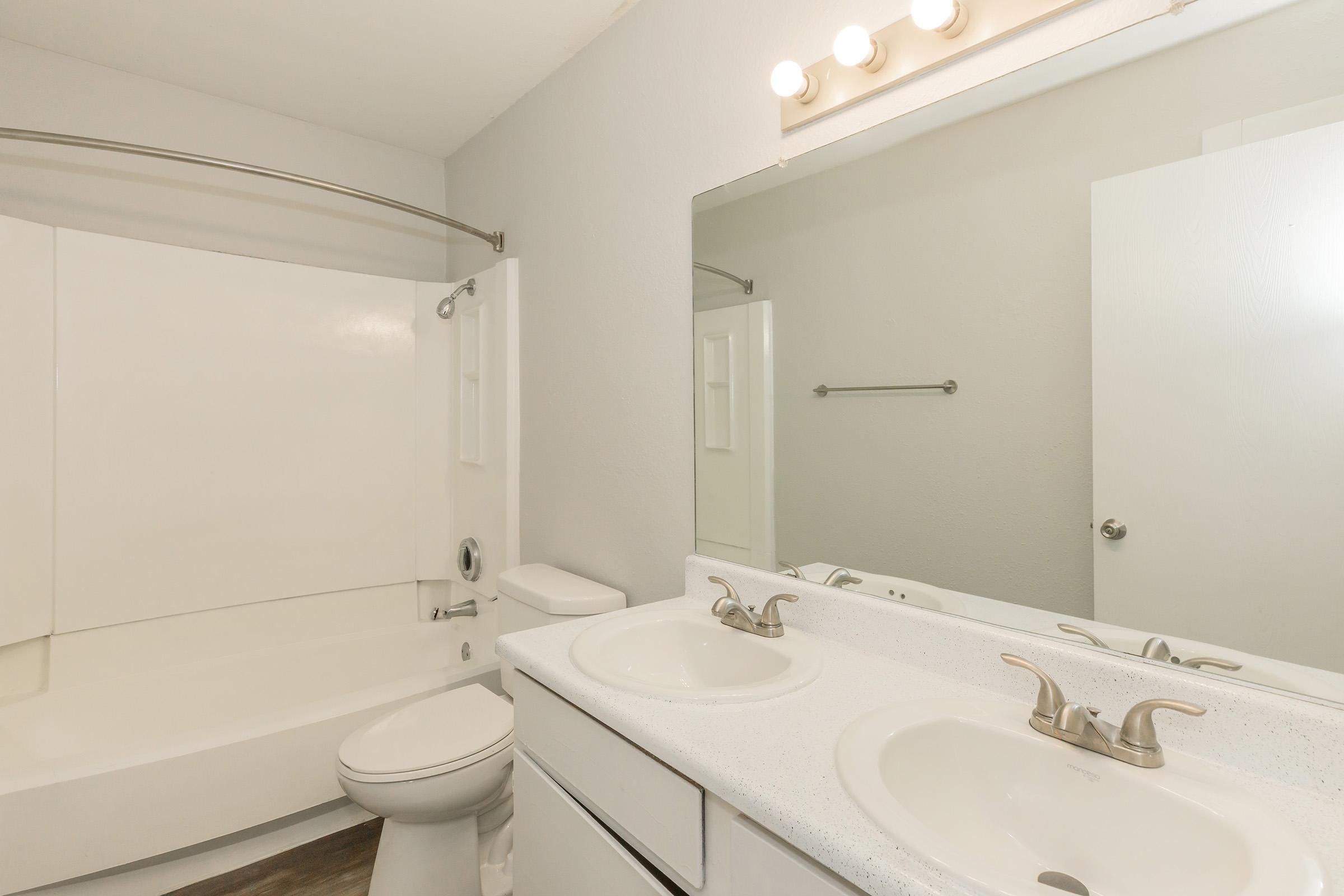 A clean, modern bathroom featuring a white tub and shower combination, a double vanity with two sinks and chrome faucets, a large mirror with overhead lights, and a light gray wall color. The flooring is a dark wood laminate, enhancing the contemporary look.