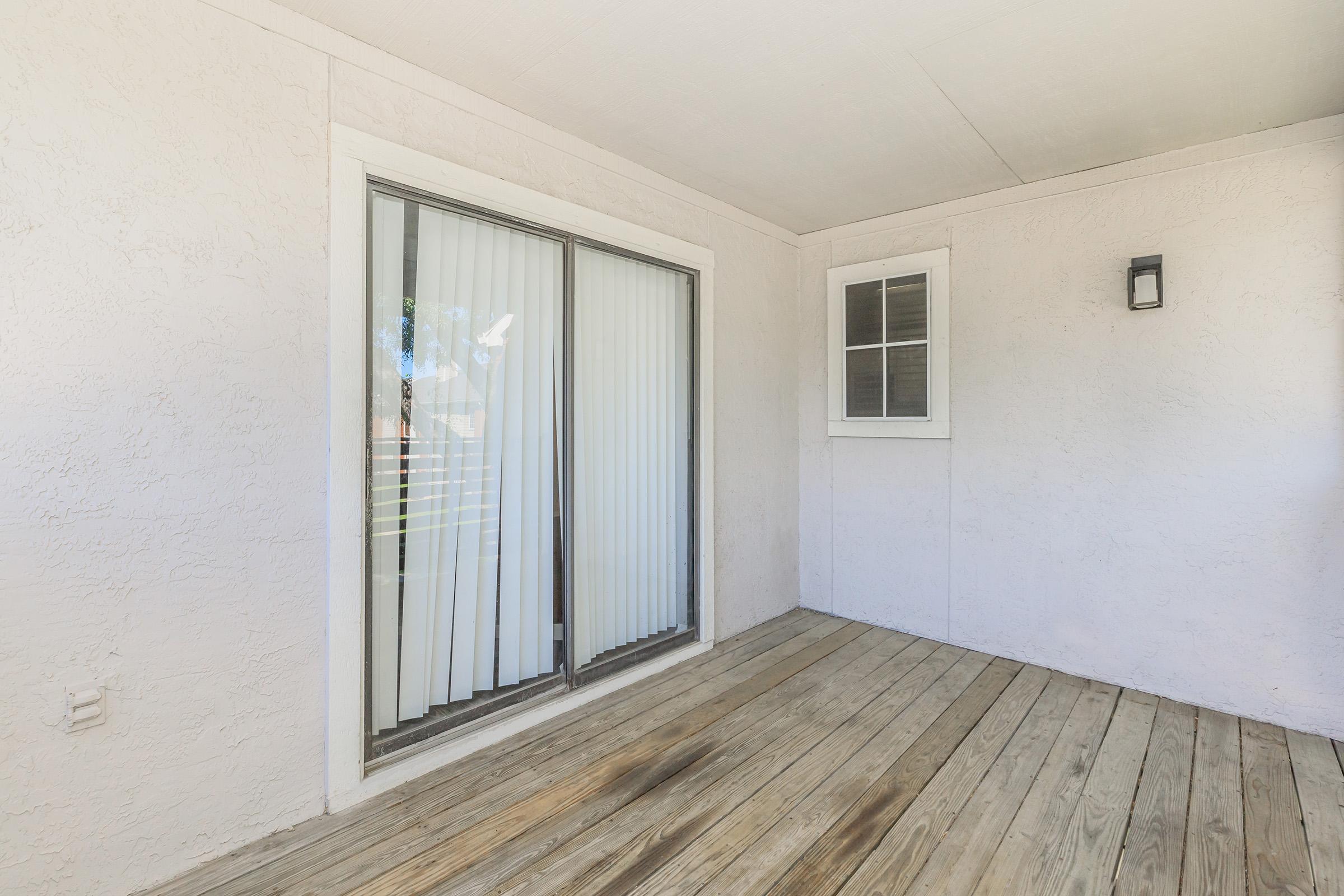 A wooden deck area with a sliding glass door and vertical blinds. There's a small window with a grid design next to the door, and the walls are painted white, giving the space a bright and airy feel.