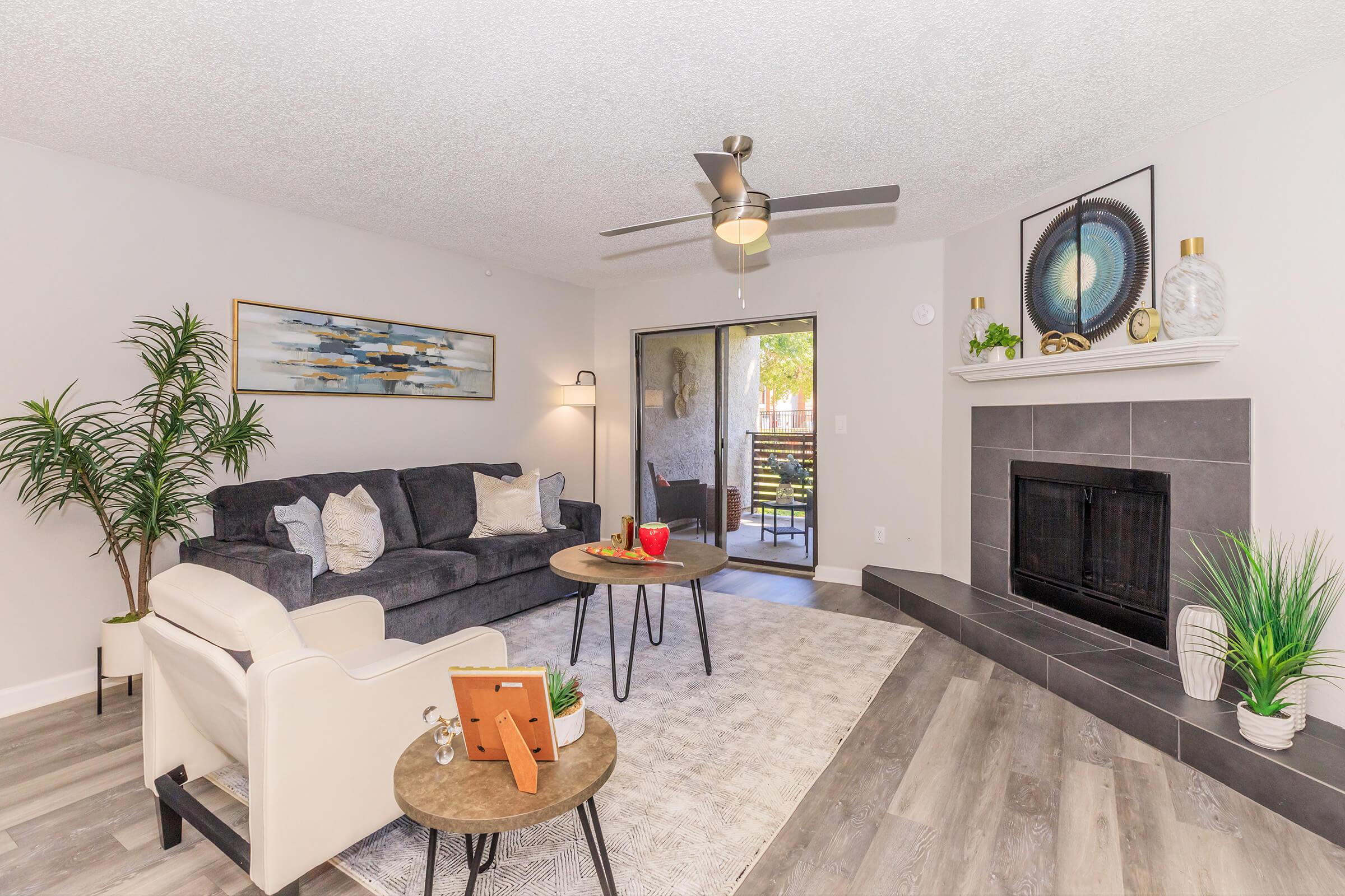 A cozy living room featuring a dark sofa, light-colored armchair, and a circular coffee table with decorative items. A ceiling fan is above, and a fireplace with a modern design is present. Large windows lead to a balcony, with green plants adding a touch of nature to the space. The room has a warm, inviting atmosphere.