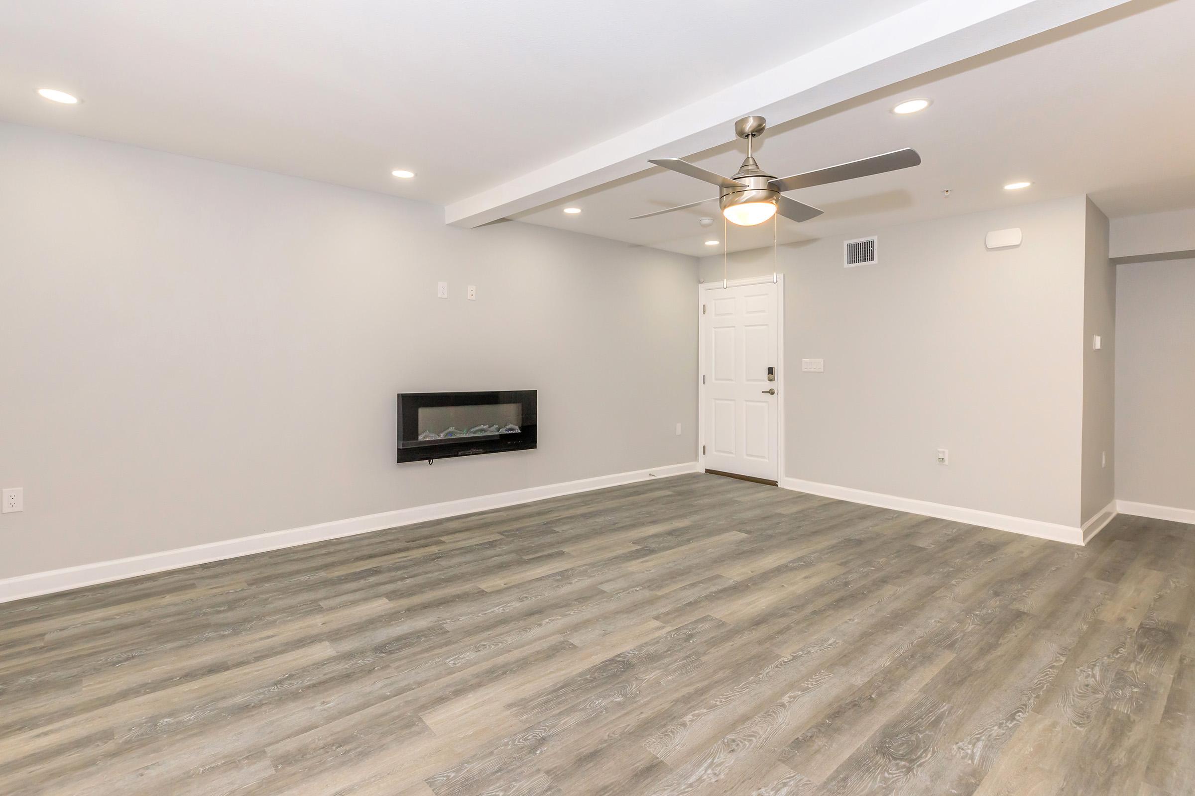 Spacious modern living room with light gray walls and hardwood-style flooring. Features an electric fireplace mounted on the wall, a ceiling fan, and a door leading outside. The area is well-lit with recessed lighting and has a minimalistic design, creating an open and inviting atmosphere.