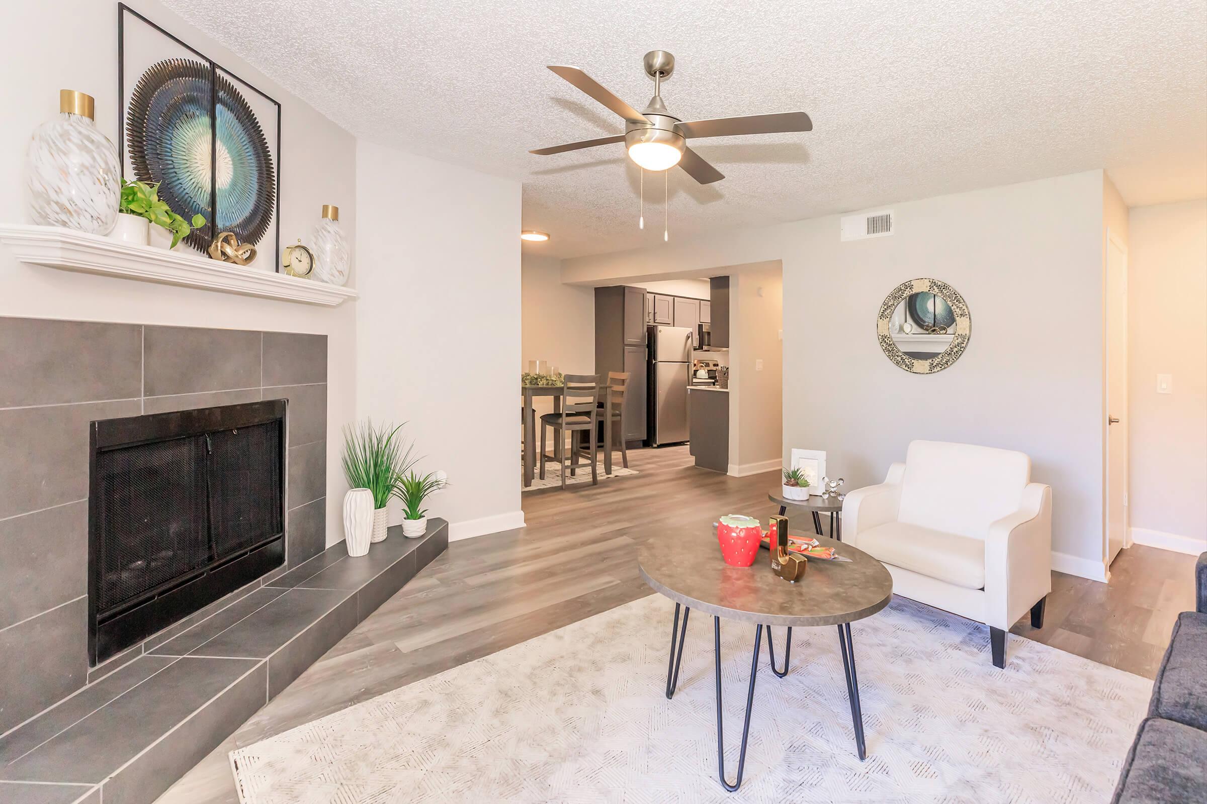 A modern living room featuring a cozy seating area with a white armchair, a round coffee table, and decorative items. There is a stylish fireplace with a gray surround, a ceiling fan, and an open kitchen visible in the background. The floor is light-colored wood, creating a warm and inviting atmosphere.