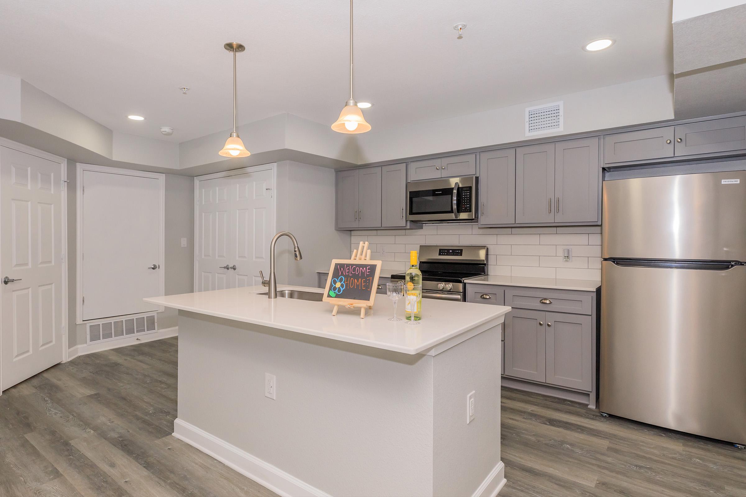 Bright modern kitchen featuring gray cabinetry, a stainless steel refrigerator and oven, and white countertops. The island includes a sink and a decorative sign saying "Welcome Home." Soft lighting fixtures hang above, and the flooring is a light, wood-like vinyl. A welcoming and spacious atmosphere.