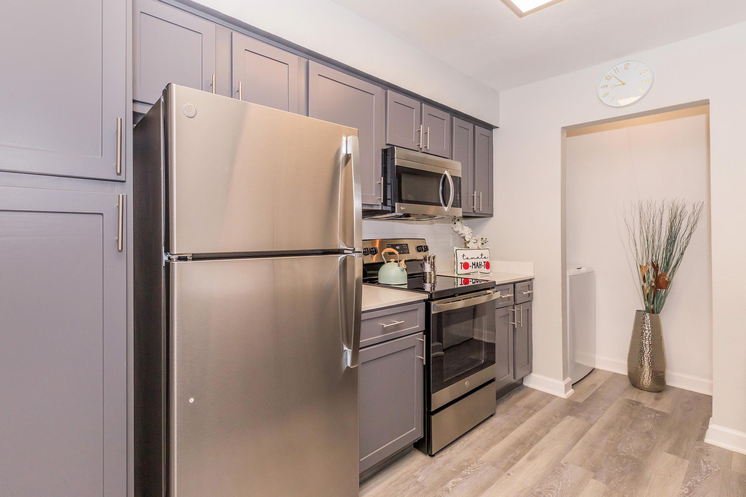 Modern kitchen featuring stainless steel appliances, including a refrigerator and microwave. The cabinetry is a dark gray with a sleek design. A small clock hangs on the wall, and a decorative vase with plant elements adds a touch of greenery. The flooring is a light wood look, enhancing the contemporary feel.