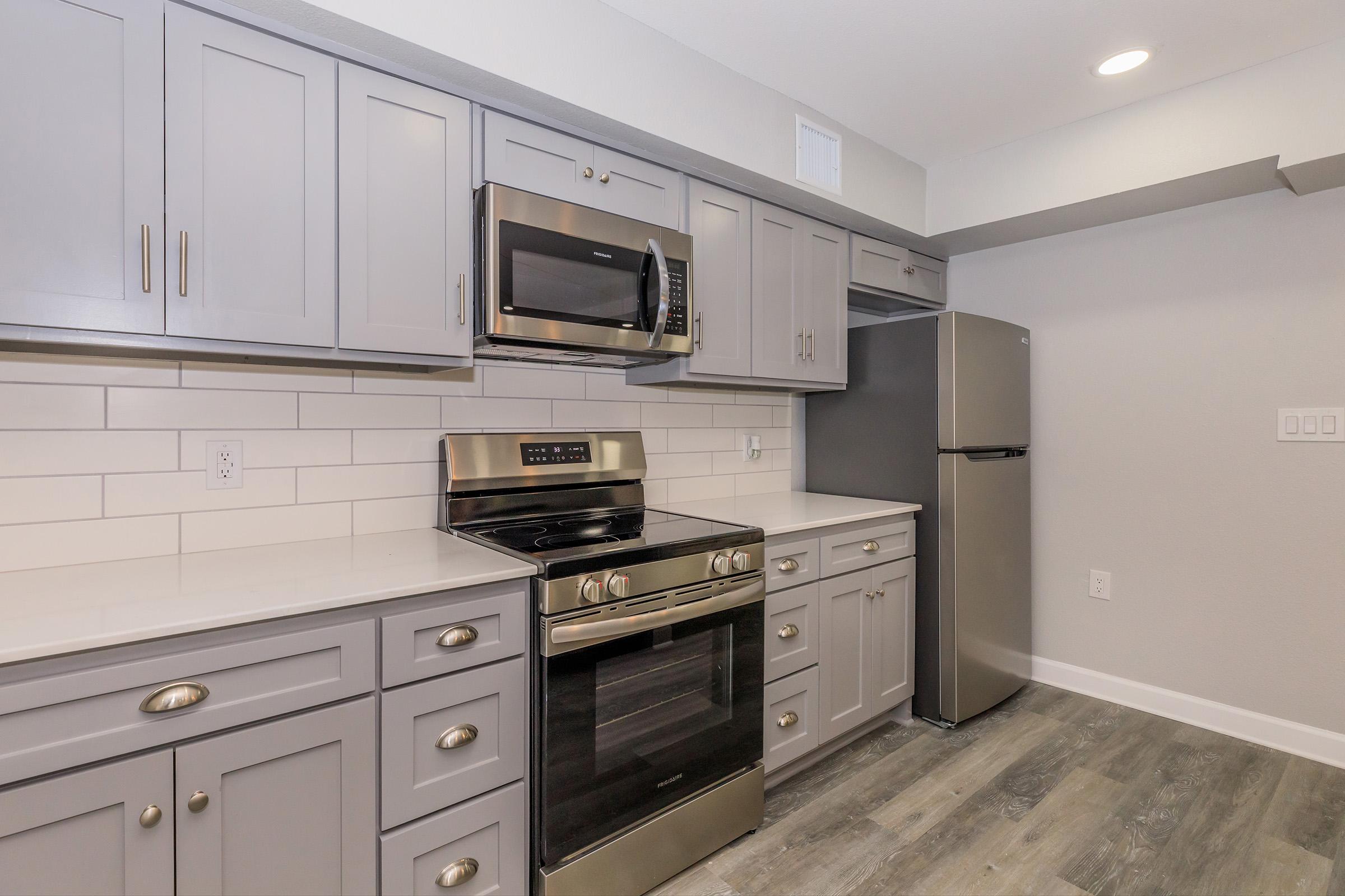 Modern kitchen featuring gray cabinetry, stainless steel appliances including an oven, microwave, and refrigerator, and a white subway tile backsplash. The space has a light-colored floor and recessed lighting, creating a bright and inviting atmosphere.