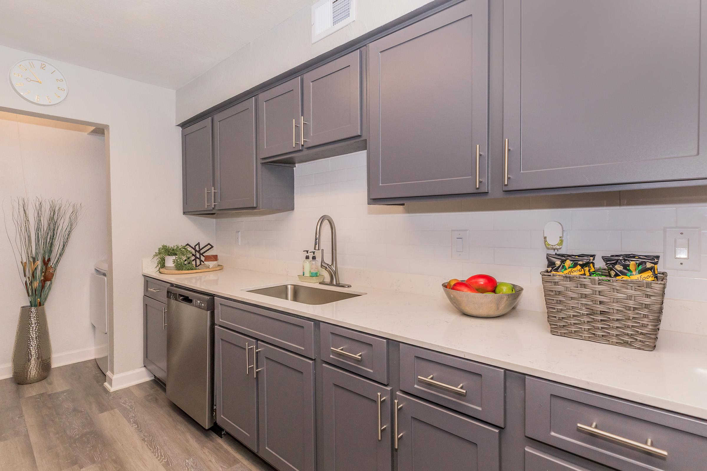 A modern kitchen featuring dark gray cabinetry, a stainless steel dishwasher, and a sleek sink. The countertop is white with a subtle pattern. A bowl of red and green apples sits next to a wicker basket filled with snacks. A decorative plant adds a touch of greenery, and a wall clock is visible in the background.