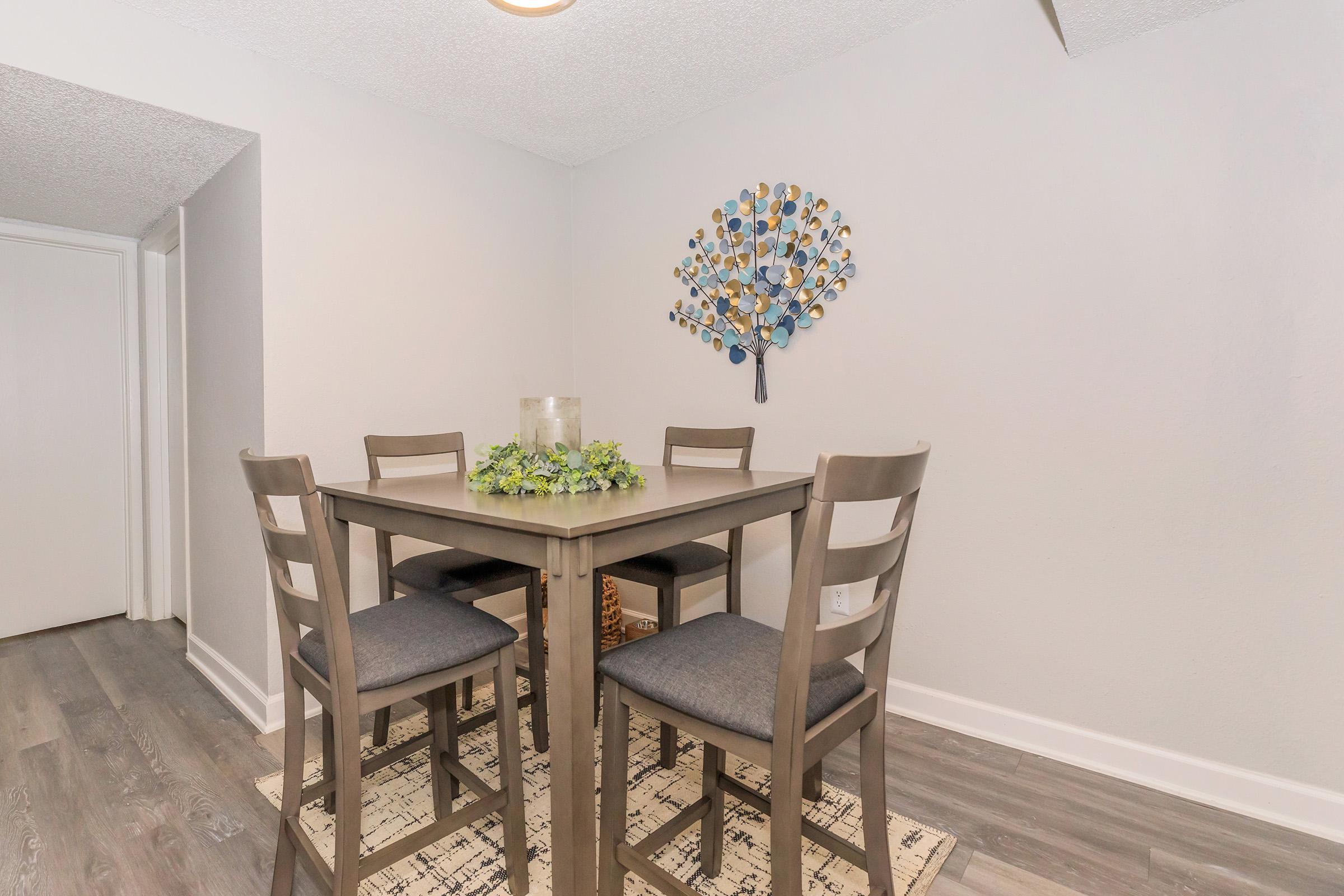 A small dining area featuring a square wooden table surrounded by four chairs with cushions. A decorative tree artwork hangs on the wall, and a centerpiece with greenery is placed on the table. The room has light gray walls and a light-colored floor, creating a cozy and modern atmosphere.