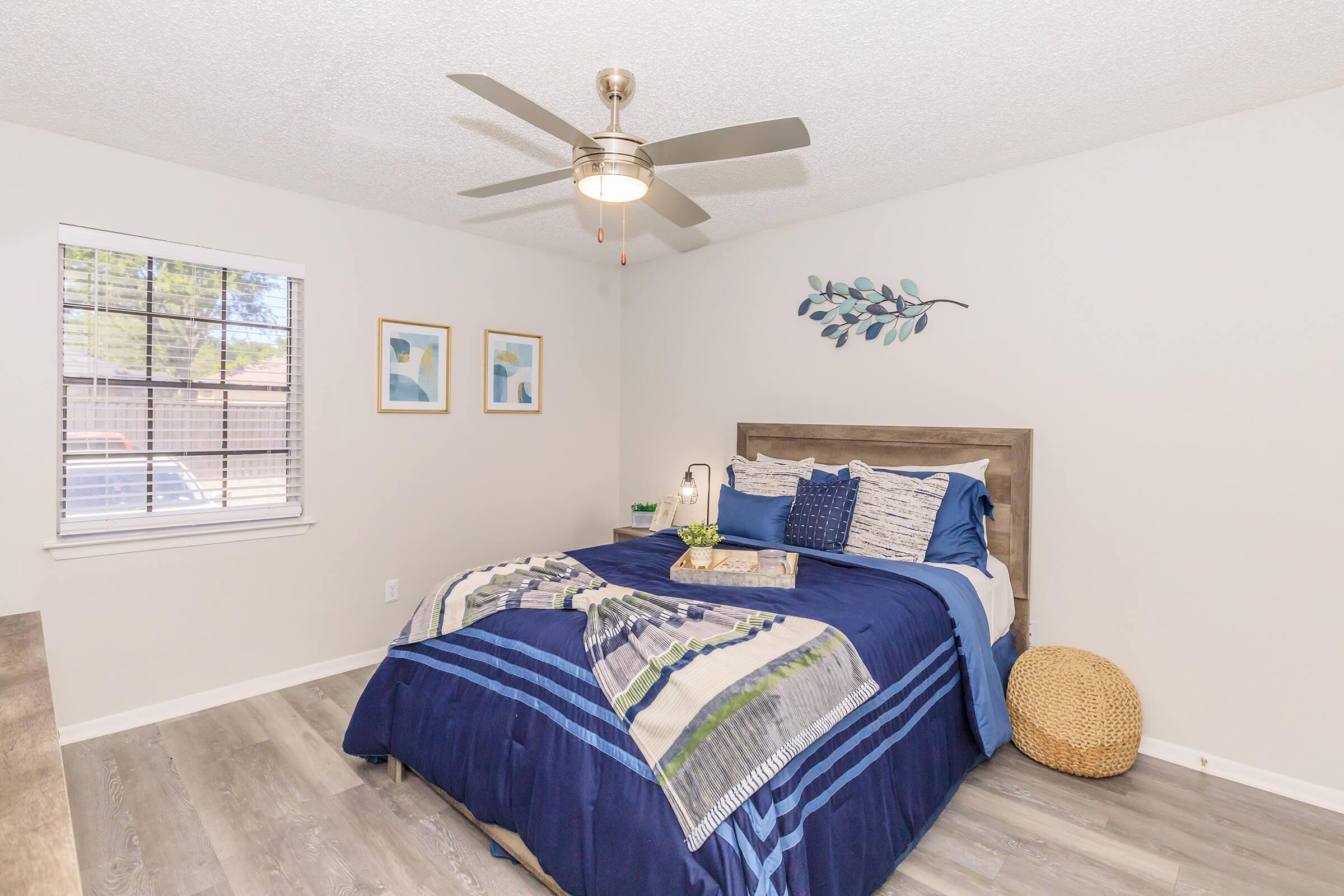 A cozy bedroom featuring a neatly made bed with a blue and white patterned bedspread, decorative pillows, and a wooden headboard. A ceiling fan hangs above, and there are two framed artworks on the wall. A small side table holds a potted plant, and a wicker basket is placed on the floor beside the bed. Natural light comes through a window with blinds.