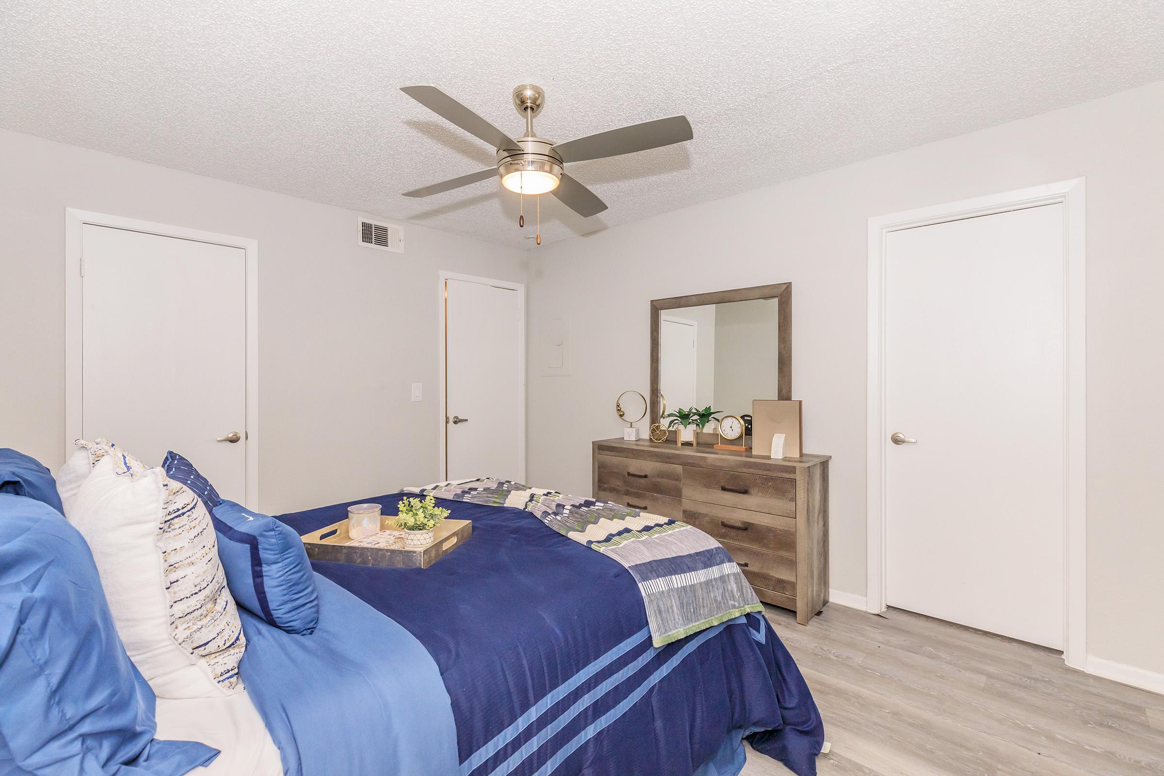 A modern bedroom featuring a blue and white bedspread with accent pillows, a wooden dresser with decor items, a mirror, and a ceiling fan. The walls are painted light gray, and there are two white doors, adding to the contemporary decor. Natural light is softly illuminating the space.