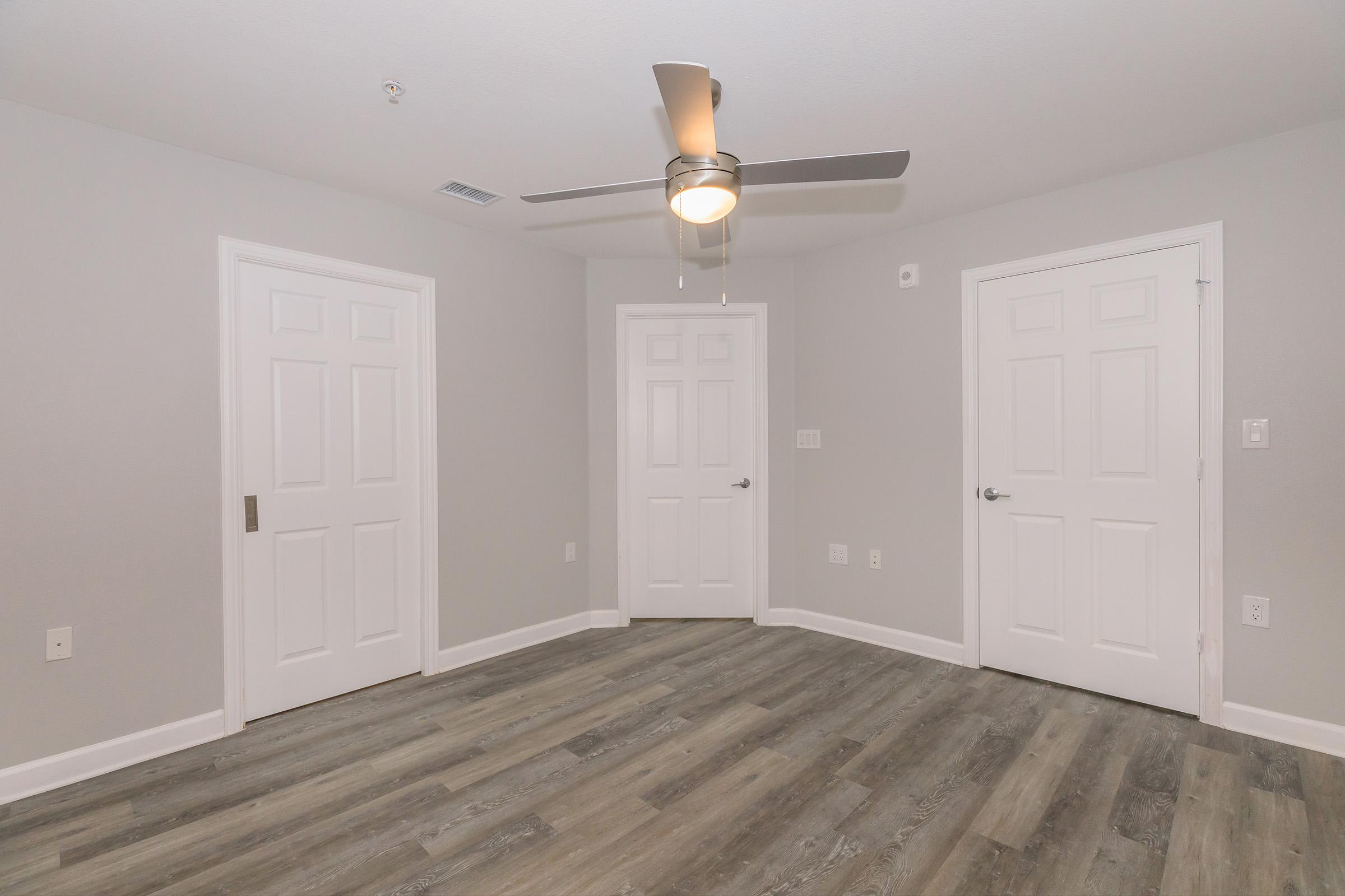 An empty room with a light-colored wall and laminate flooring. The space features a ceiling fan and two closed white doors on opposite walls, providing a simple and clean aesthetic. The room is well-lit and appears spacious, suitable for various uses.
