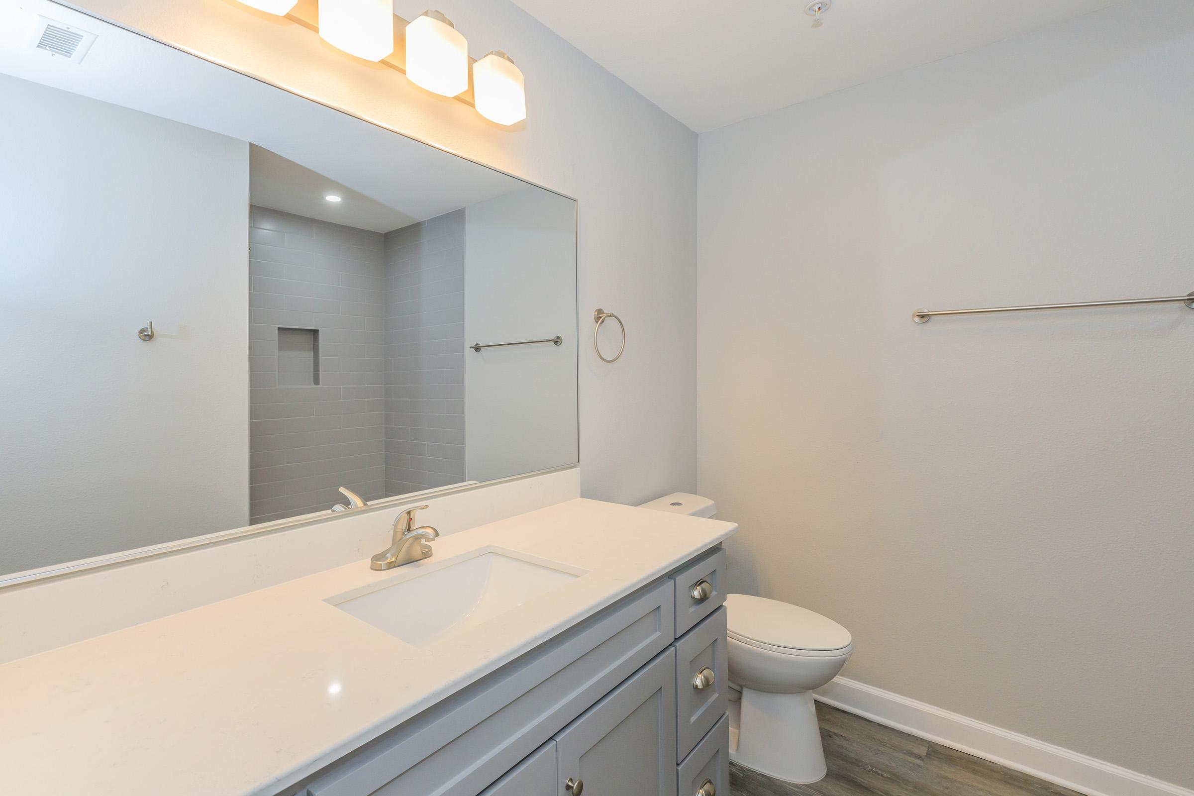 A modern bathroom featuring a gray vanity with a white countertop, a large mirror, and three light fixtures above. There is a toilet and a towel bar on the wall, with a shower area visible in the background. The walls are painted light gray, and the flooring has a wood-like appearance.