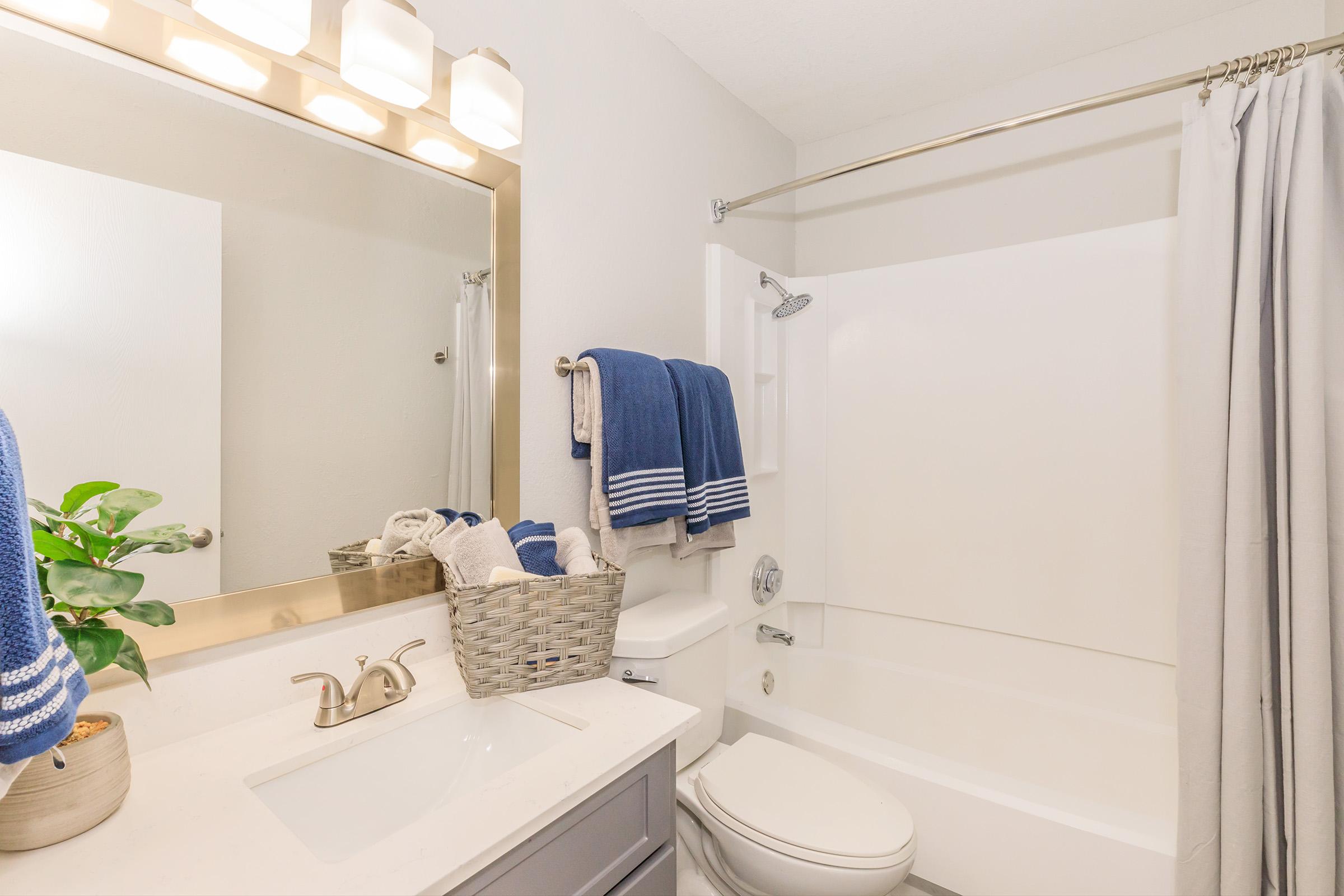 A clean, modern bathroom featuring a white bathtub, a stylish mirror with lights above, and a vanity with a sink. Blue towels hang neatly, and a woven basket sits on the countertop. A plant adds a touch of greenery, and soft gray curtains frame the shower area, creating a welcoming atmosphere.