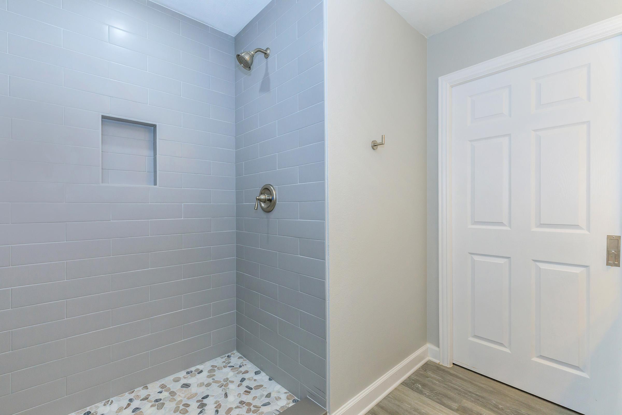 Modern bathroom featuring a walk-in shower with gray tiles and a built-in niche. The shower has a round showerhead, and a decorative pebble-style tile floor. A white door is visible, leading out of the bathroom, and there is a single wall hook beside the shower.