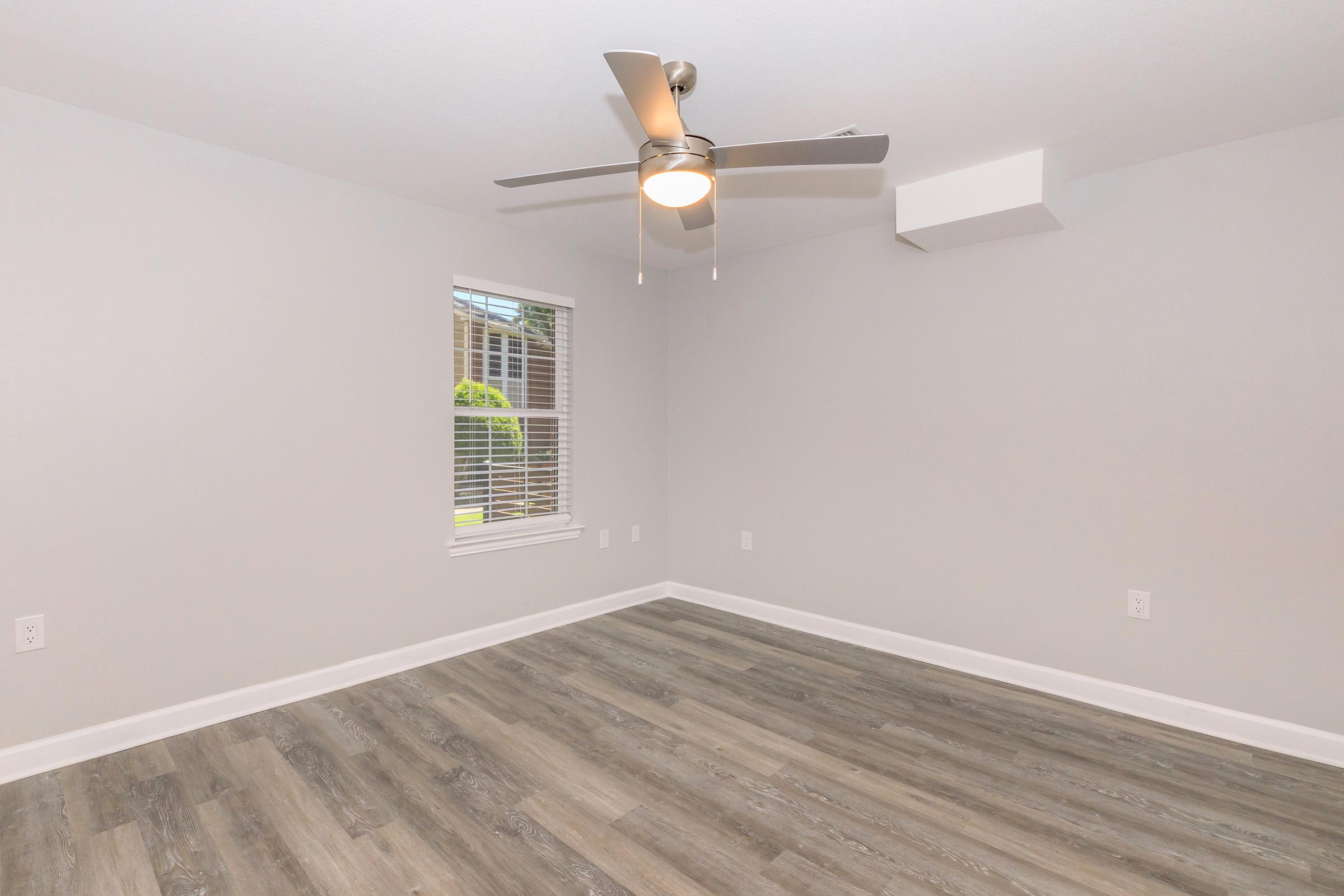 A vacant room with light gray walls and a wooden floor. There is a ceiling fan with a light fixture in the center. A window with white blinds allows natural light into the space, and there is a vent on the wall. The room is unfurnished, creating a clean and open atmosphere.