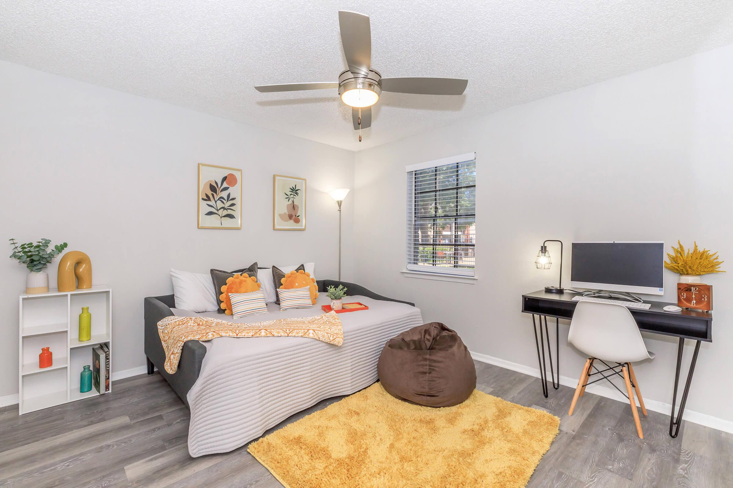 A cozy living space featuring a gray sofa adorned with decorative pillows, a small desk with a computer, and a stylish chair. The walls display framed artwork, and a floor lamp provides light. A plush rug and bean bag add comfort to the room, which has a bright window letting in natural light.