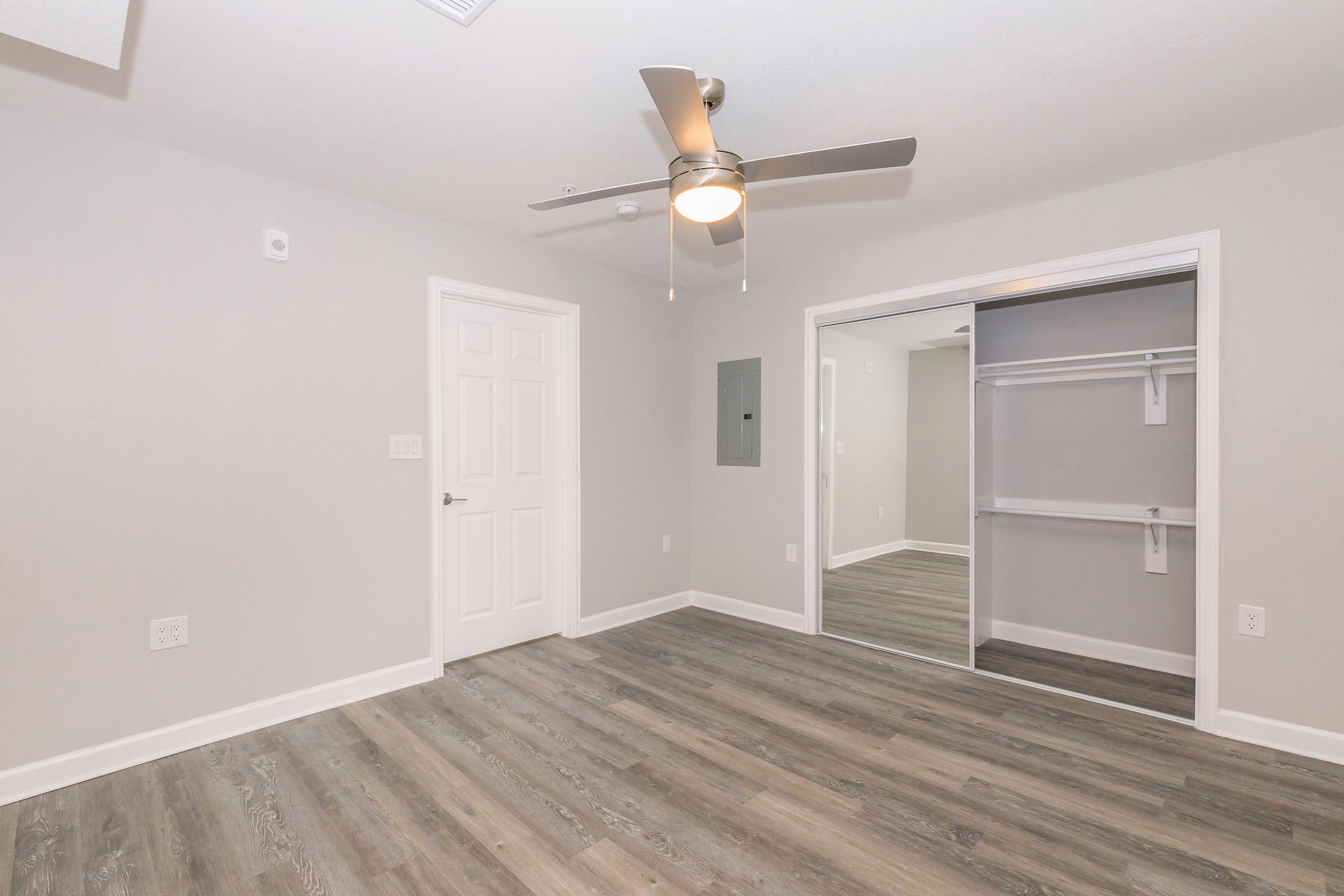 A well-lit, modern bedroom featuring light gray walls and a ceiling fan. The room has a large mirror closet with white shelving and doors, and light-colored wood flooring. There is a white door on one side and an electrical panel on the wall. The overall ambiance is clean and minimalistic.