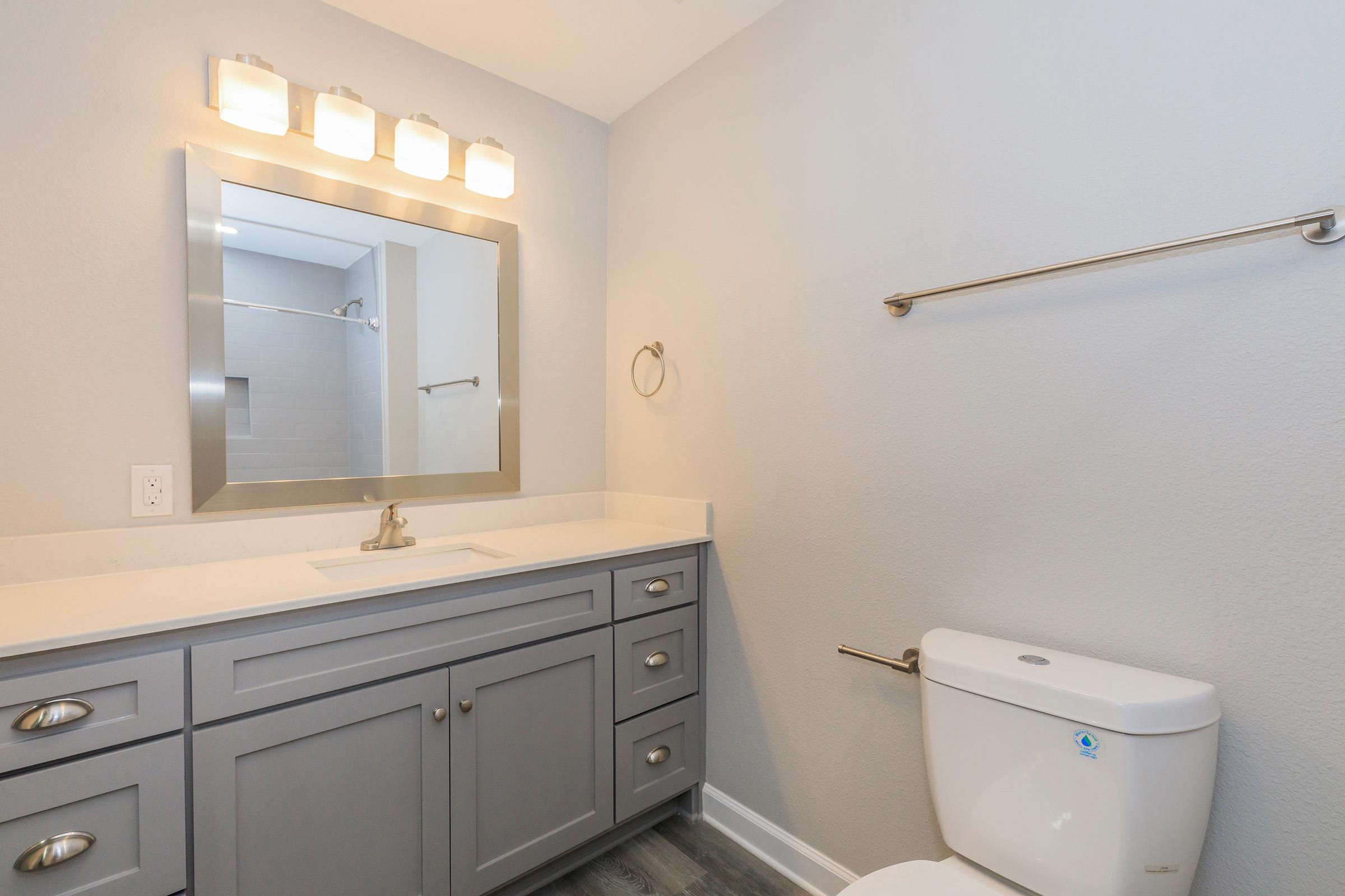 Modern bathroom featuring a gray vanity with a white countertop, a large mirror with four light fixtures above, a towel rack, a toilet, and gray flooring. The walls are painted in a light color, creating a bright and spacious atmosphere.