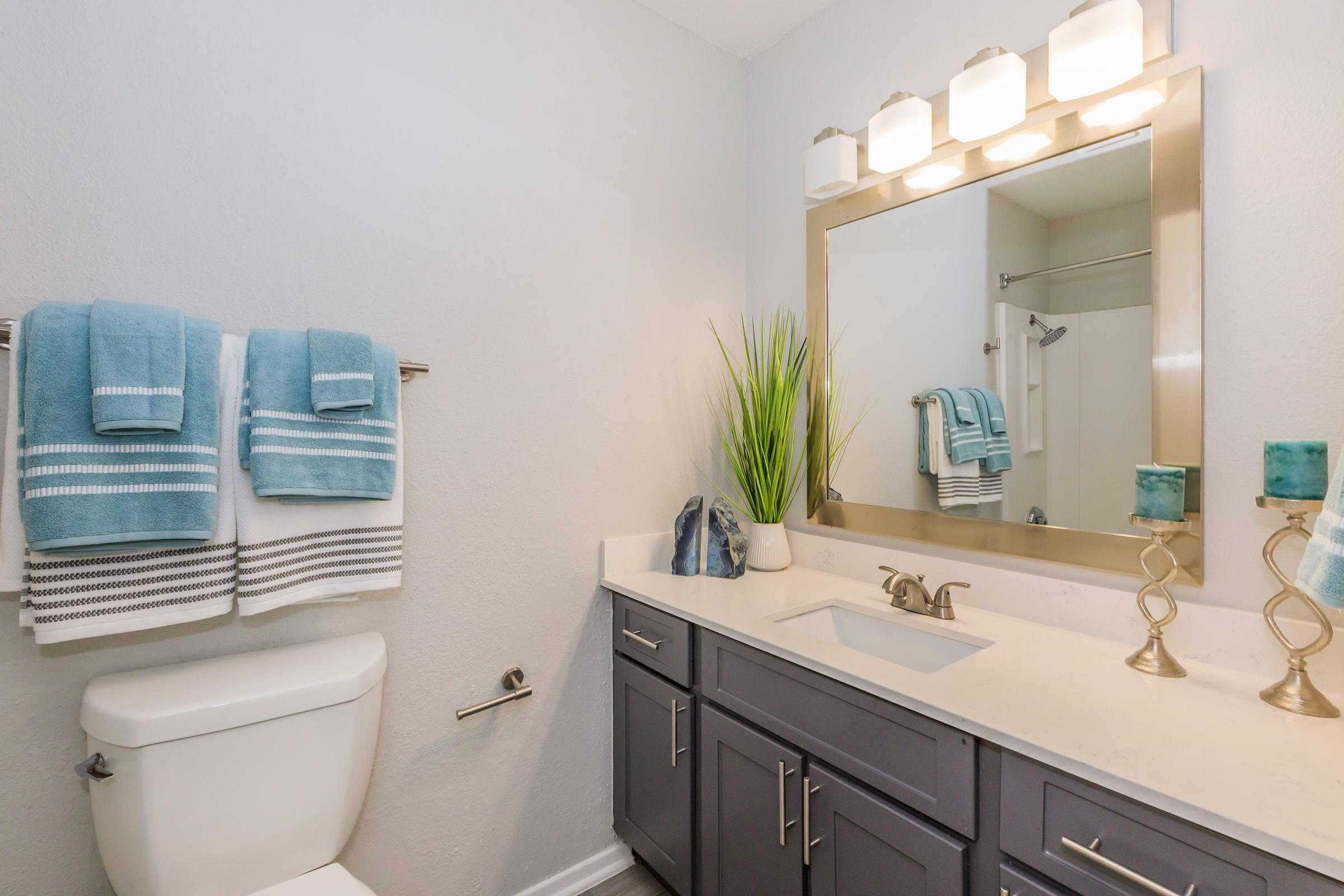 A modern bathroom featuring a gray vanity with a white countertop, a large mirror with decorative lighting, a toilet, and neatly arranged towels in aqua and white. A potted plant adds a touch of greenery, while decorative candle holders sit on the counter, creating a clean and inviting atmosphere.