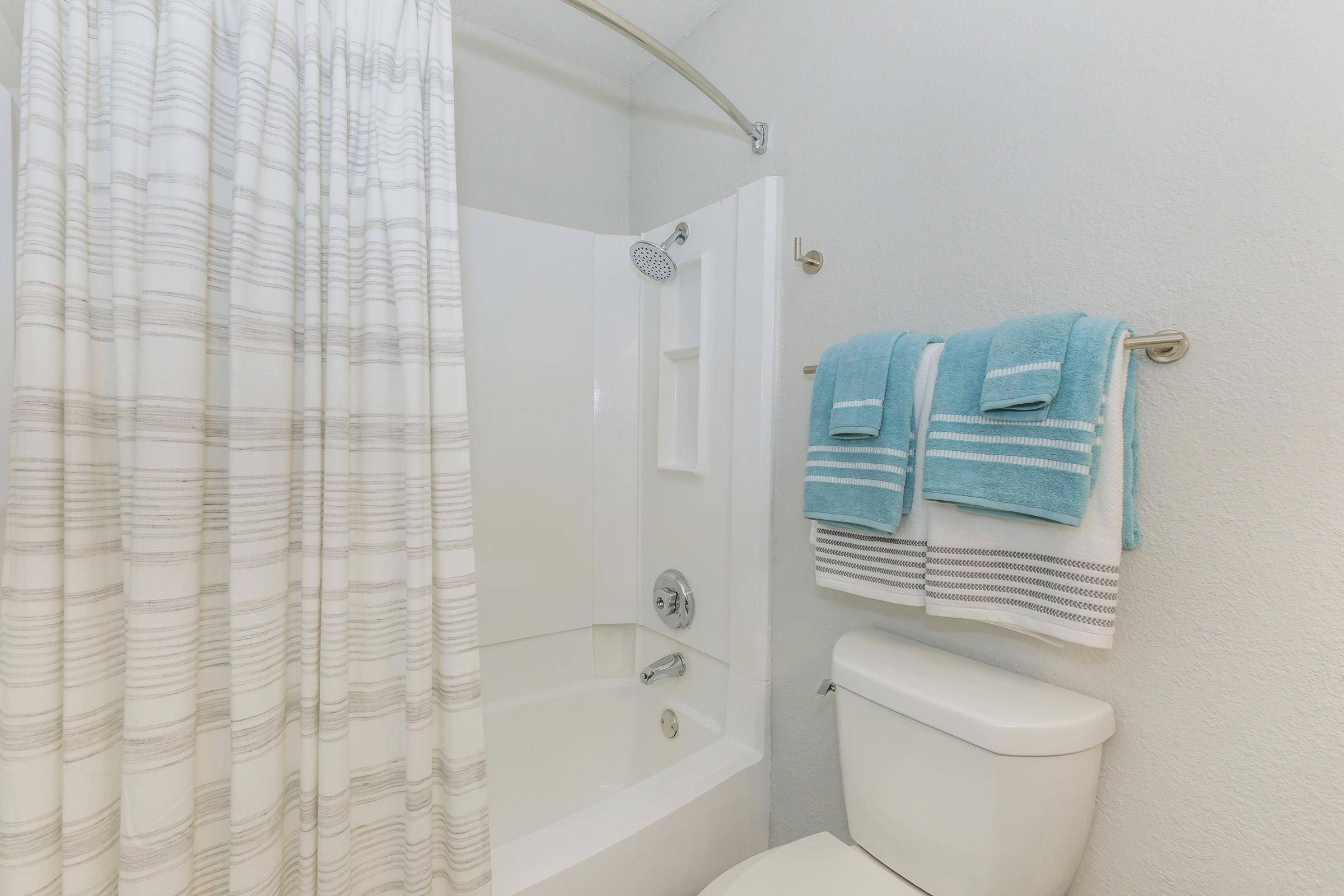 A clean, modern bathroom featuring a white bathtub with a curved shower curtain rod. The curtain has a striped pattern. A white toilet is positioned next to the bathtub, and neatly folded blue and white towels are hanging on a towel rack. The walls are painted in a light color, creating a spacious feel.