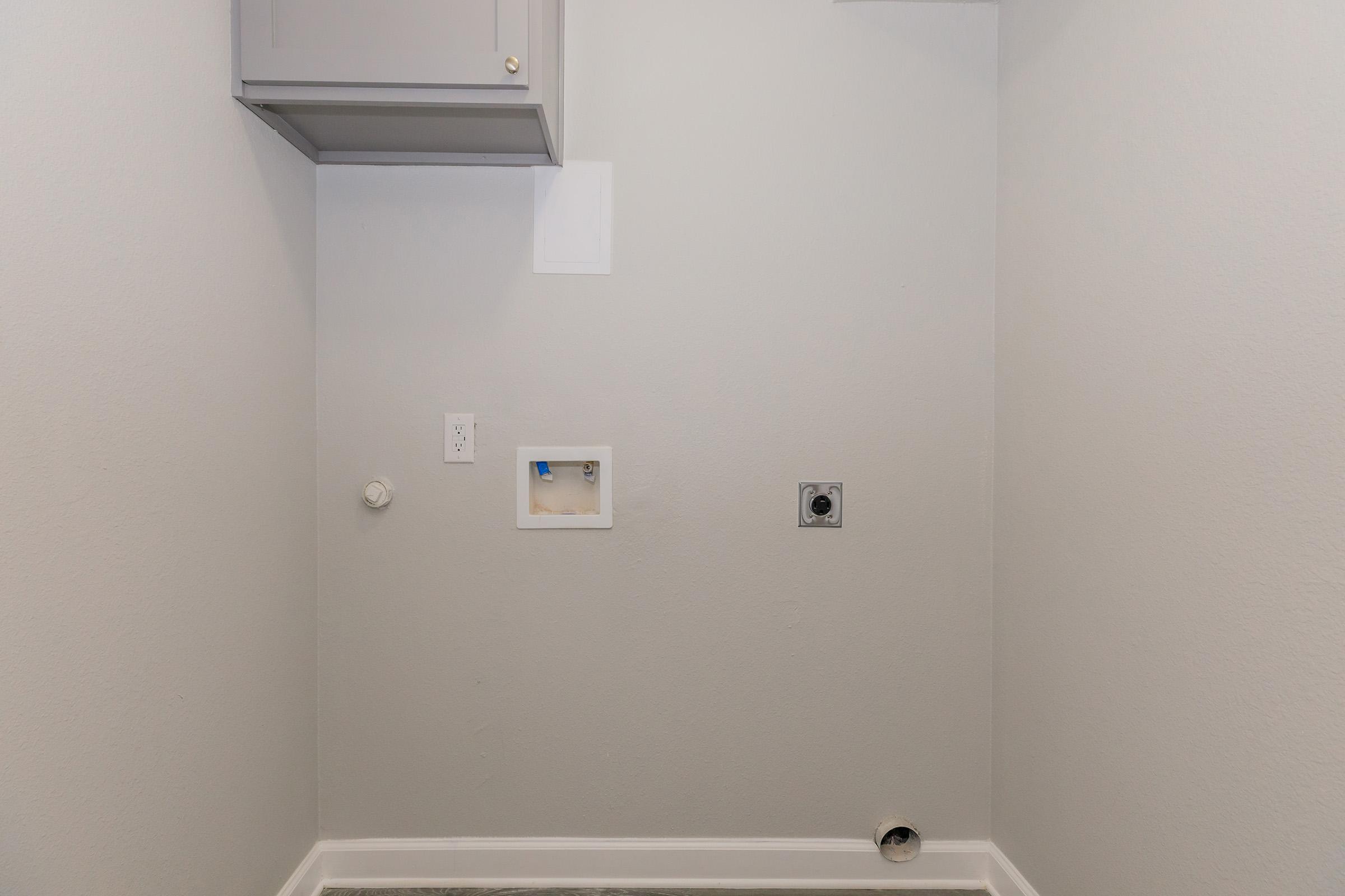 Empty laundry room with light gray walls and a small cabinet above. There are electrical outlets and a water connection on the wall, but no appliances or decor present, creating a clean and spacious look. The floor is tiled, and the space is well-lit.