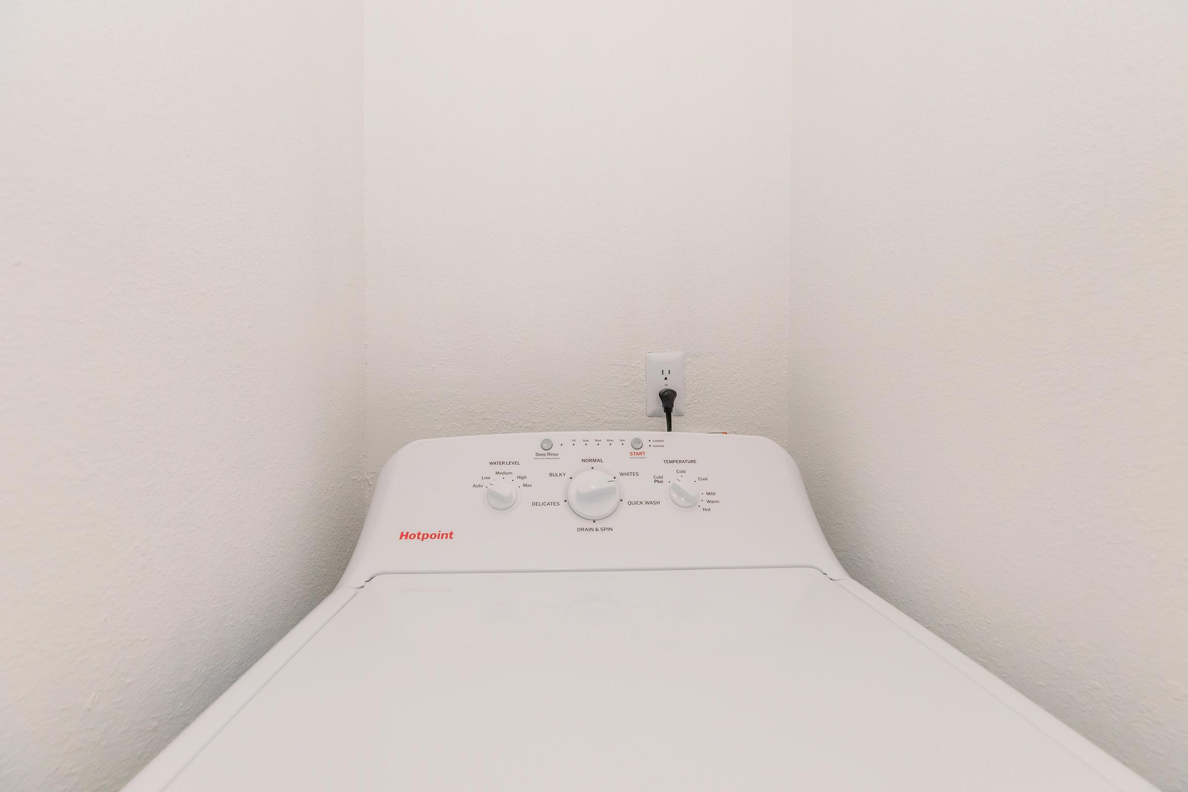 A top-loading washing machine positioned in a corner. The control panel features various dials and buttons, with a power outlet visible above it. The walls are plain and white, creating a clean and minimalistic laundry space.