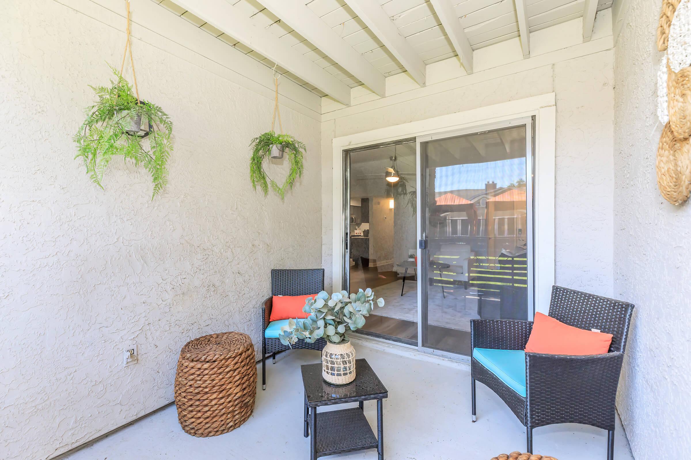A cozy outdoor patio featuring two wicker chairs with blue cushions and orange accents, a small table, and a decorative plant. Hanging planters with greenery adorn the walls, and a sliding glass door leads to the interior space, creating a welcoming atmosphere.