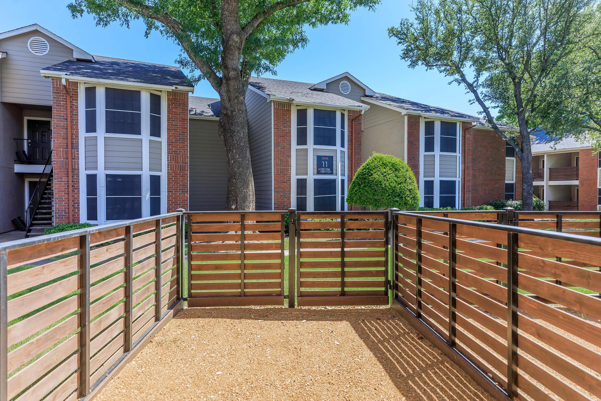 A fenced outdoor area featuring wooden slats, with gravel ground. In the background, there are two residential apartment buildings with large windows, and lush green trees providing shade. The scene is bright and sunny, reflecting a peaceful environment.