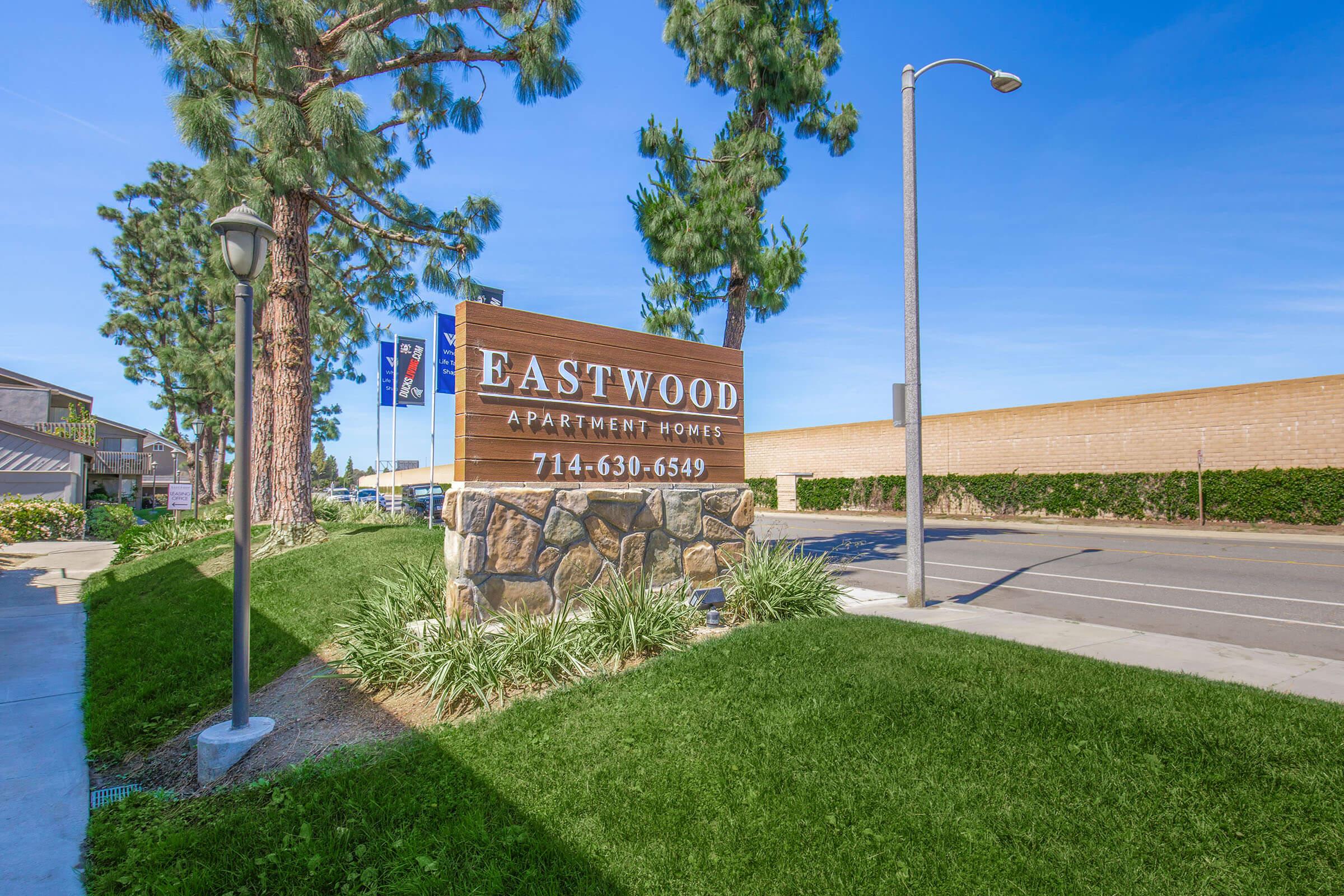 Sign for Eastwood Apartment Homes, featuring the name prominently displayed on a wooden board with stone base. Surrounding greenery includes grass and trees, with a street and light post visible in the background. A contact phone number is also included on the sign.