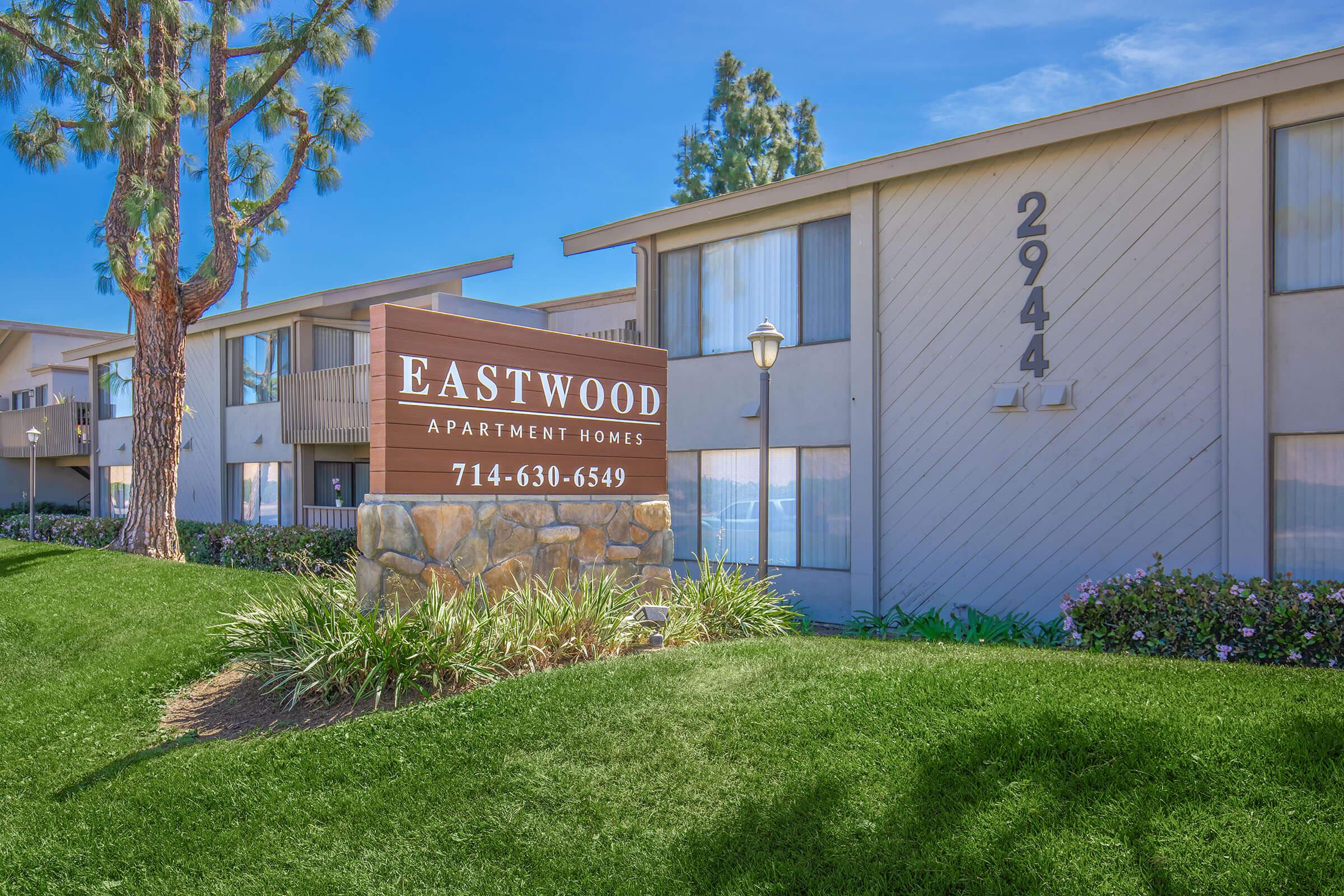 Sign for Eastwood Apartment Homes featuring the name prominently displayed. The building is two stories, with multiple windows and balconies, surrounded by green grass and shrubs. A tall tree stands nearby, and the building number "2944" is visible on the side. Clear blue sky in the background enhances the setting.