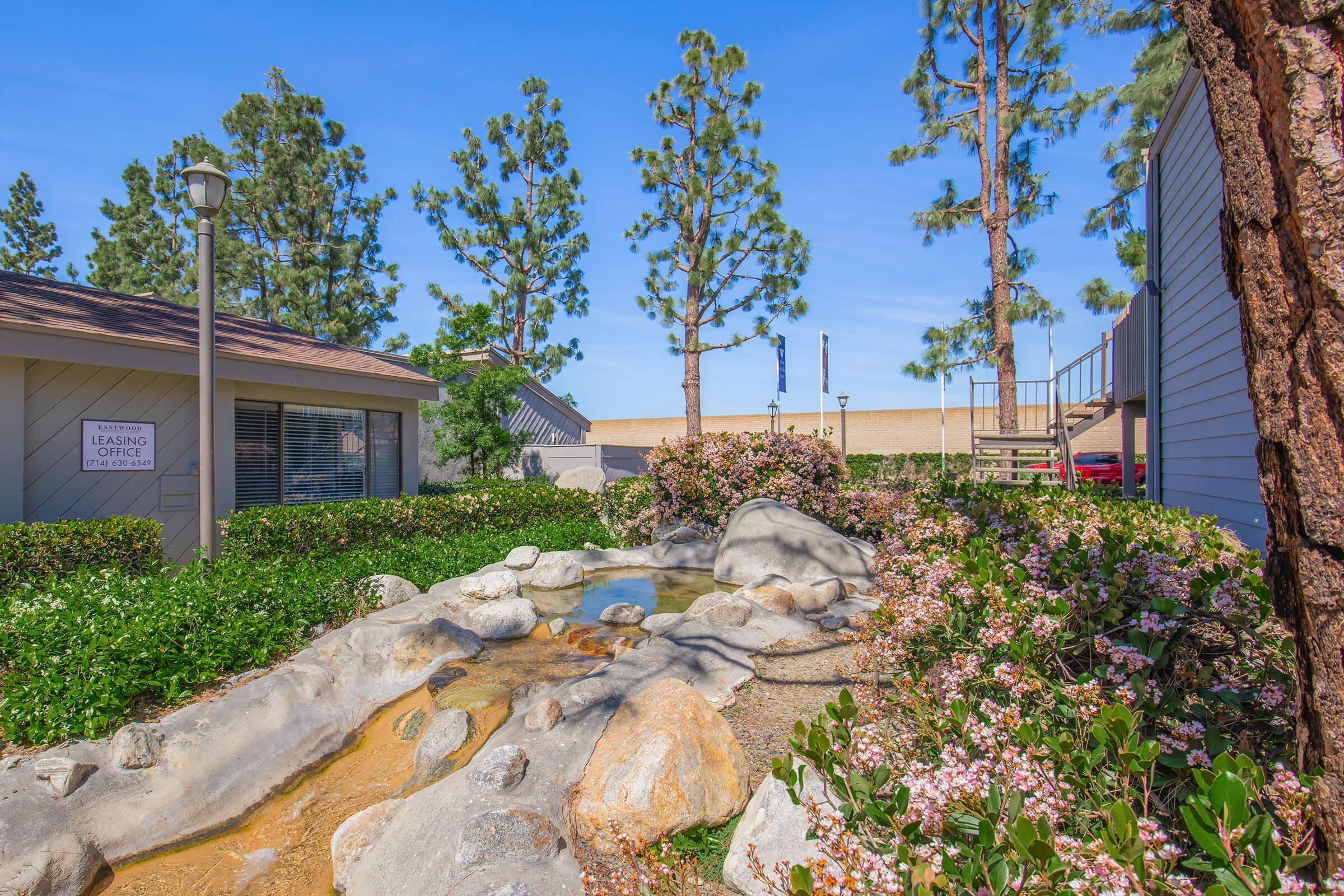 A peaceful outdoor scene featuring a landscaped area with a small stream and rocks. Colorful flowers line the path, and tall trees provide shade. In the background, a leasing office building is partially visible, set against a clear blue sky. The atmosphere is serene and inviting, ideal for relaxation.