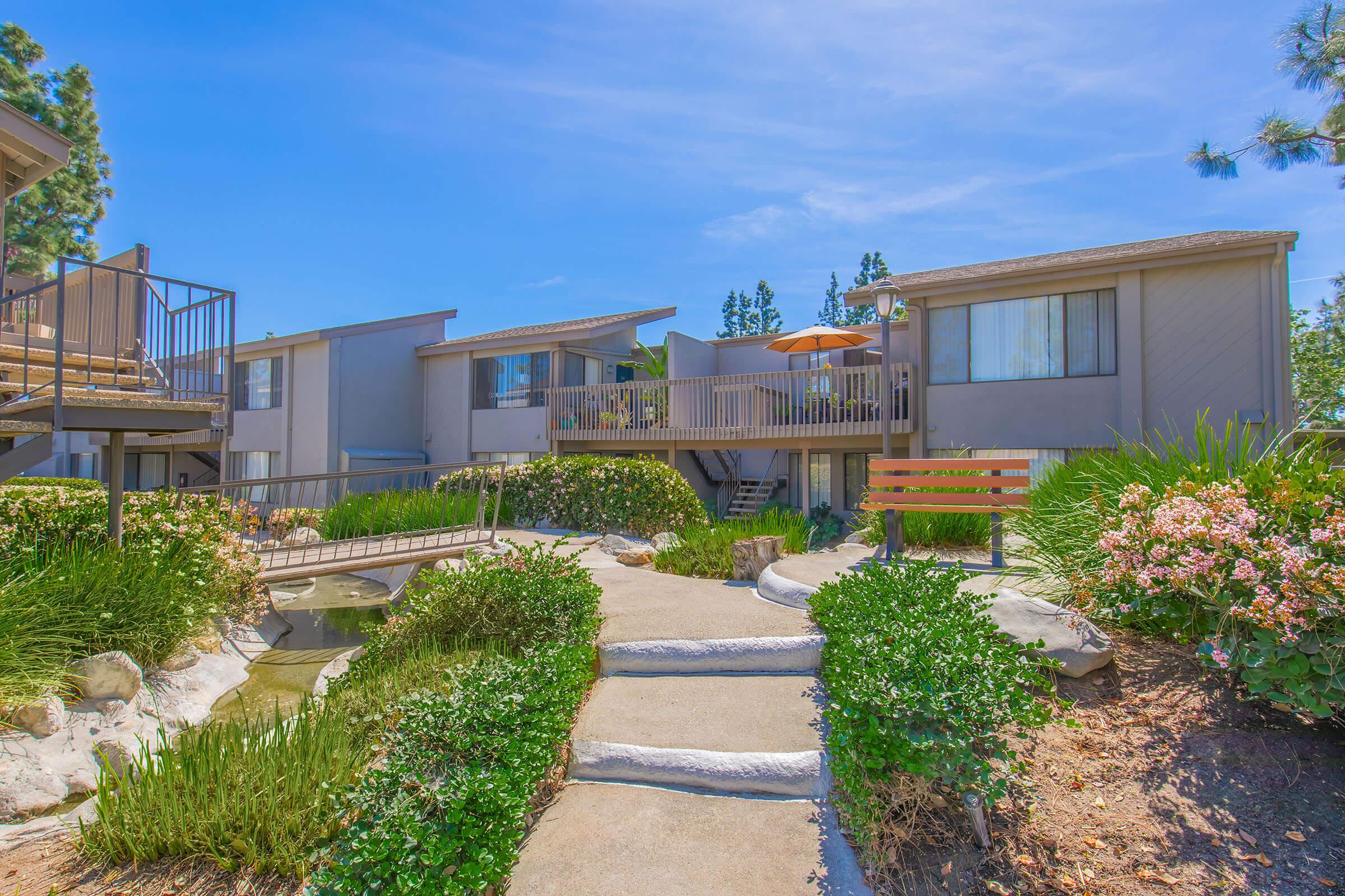 A landscaped pathway leads through greenery to a set of two-story apartment buildings, featuring balconies and outdoor seating. The sky is blue with a few clouds, and there is a small bridge over a water feature visible in the foreground. The environment appears sunny and well-maintained.
