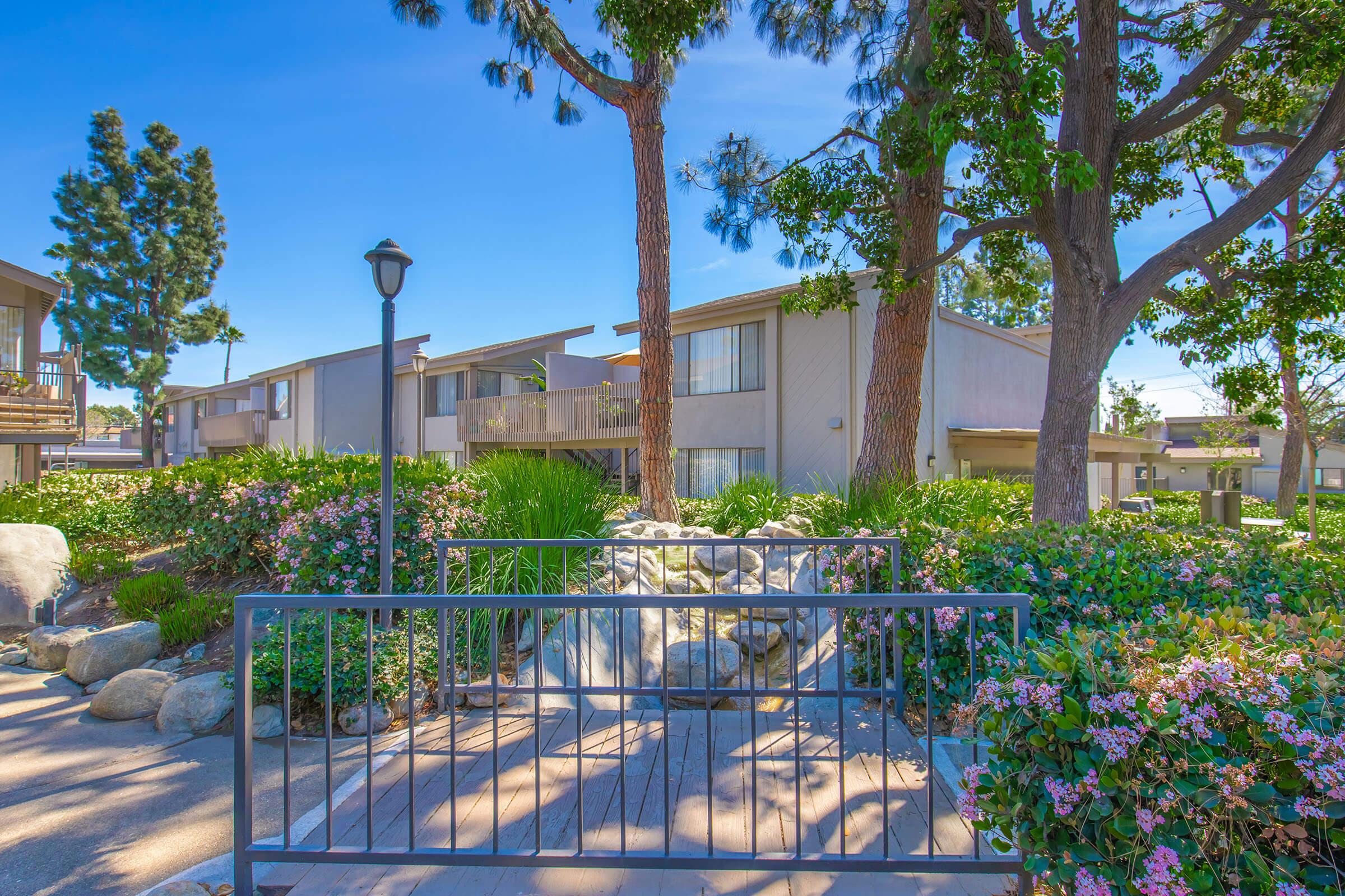 A well-maintained residential complex with landscaped pathways, featuring lush greenery and flowering plants. There are stone decorations and trees nearby, framed by a decorative gate leading to an inviting walkway. The backdrop includes two apartment buildings under a clear blue sky.