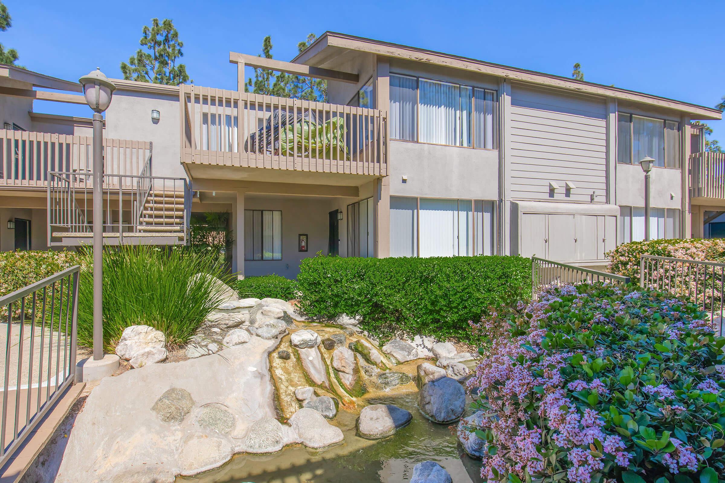 A modern two-story apartment building with a landscaped courtyard featuring a rock fountain and colorful flowers. The exterior has large windows, wooden balconies, and well-maintained greenery, creating a serene living environment in a sunny setting.