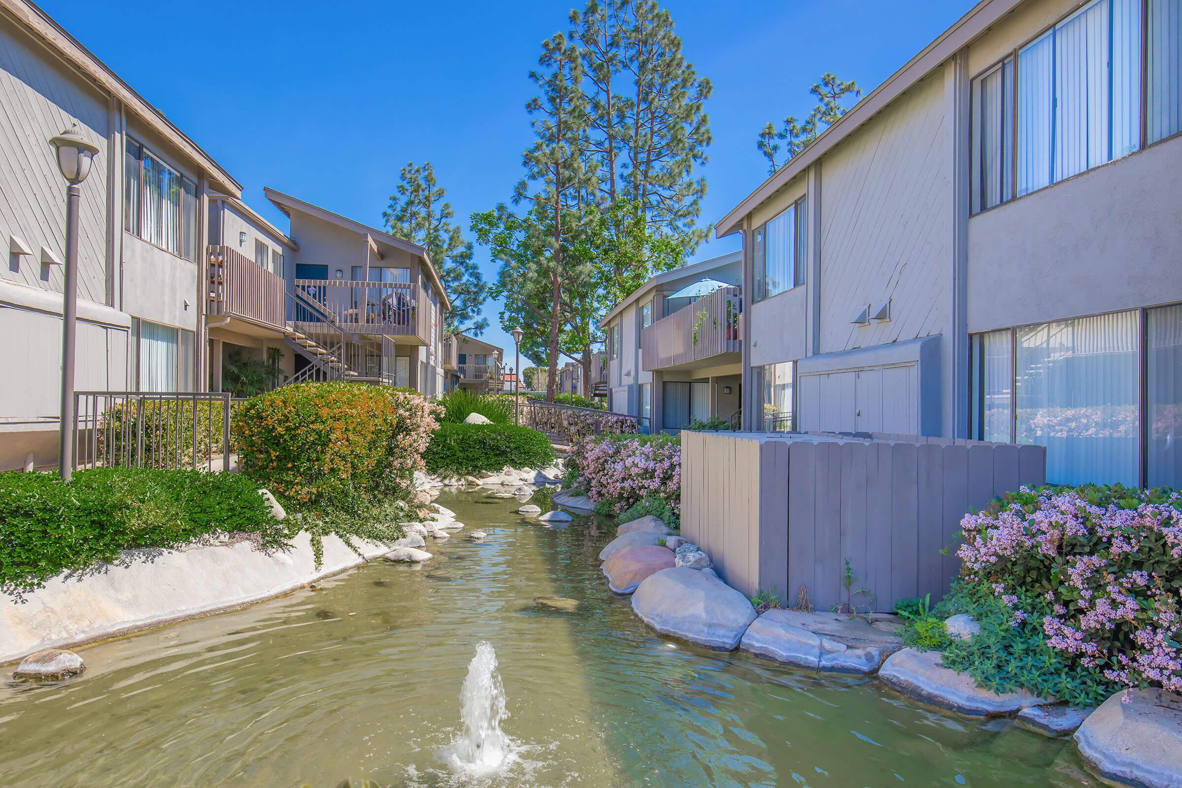 A lush apartment complex with two-story buildings flanking a serene waterway. The scene features a small fountain in the water, surrounded by greenery and colorful flowers. Tall trees rise in the background under a clear blue sky, creating a tranquil and inviting environment.