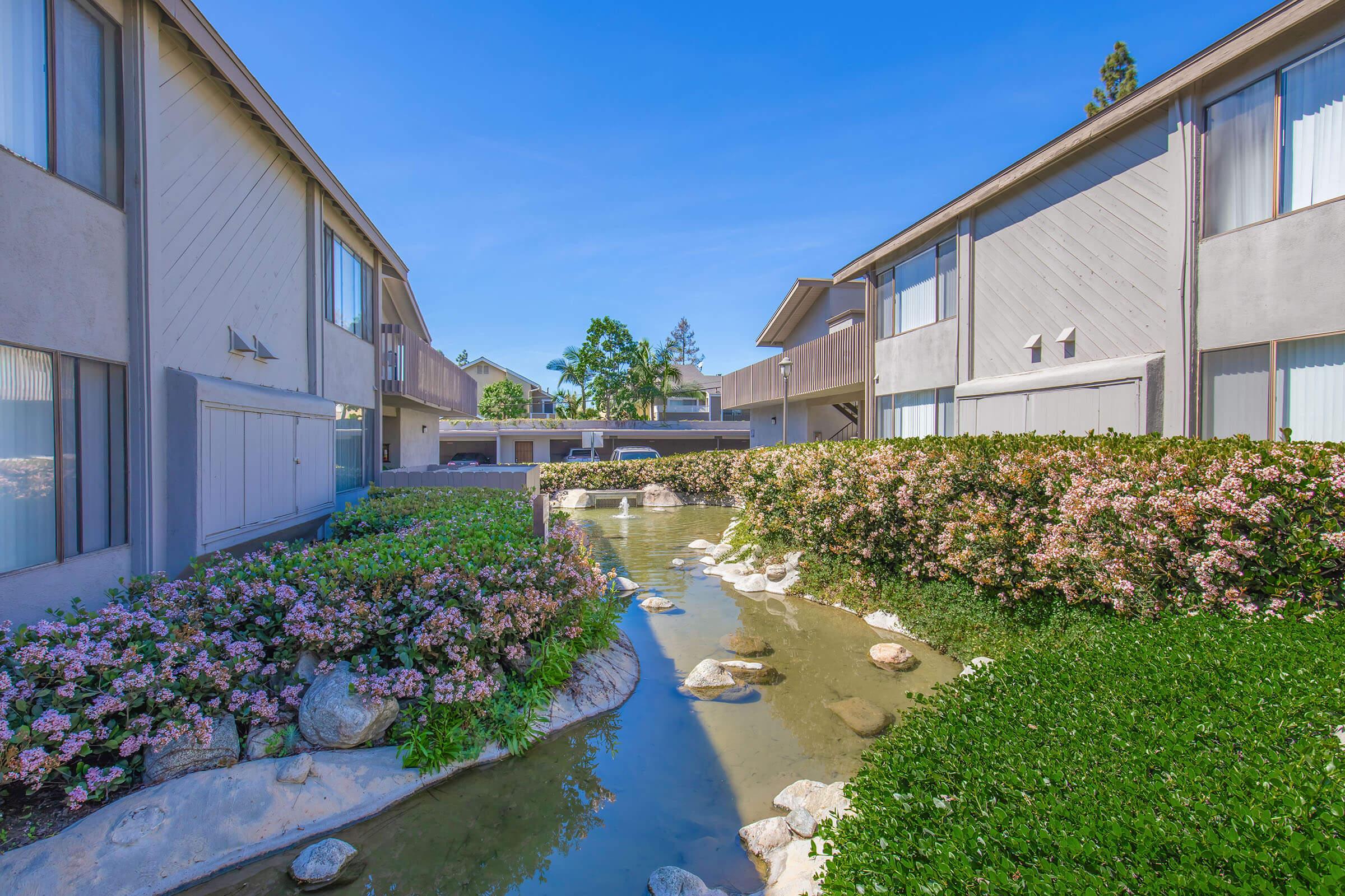 A peaceful courtyard view between two apartment buildings, featuring a meandering stream bordered by colorful flowering bushes and greenery under a bright blue sky. The setting conveys a calm and inviting atmosphere.