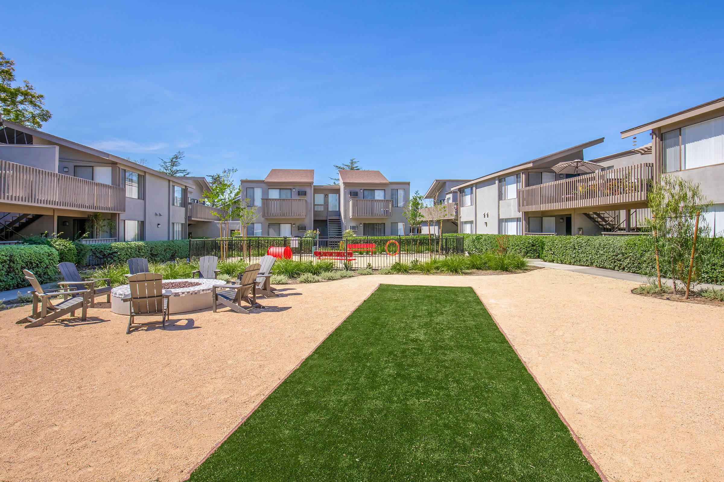 A courtyard view of an apartment complex featuring two-story buildings with balconies. There is a landscaped area with green grass and a sandy path, surrounded by trees and shrubs. Outdoor seating is available with chairs and a fire pit. Play equipment is visible in the background, suggesting a family-friendly environment.