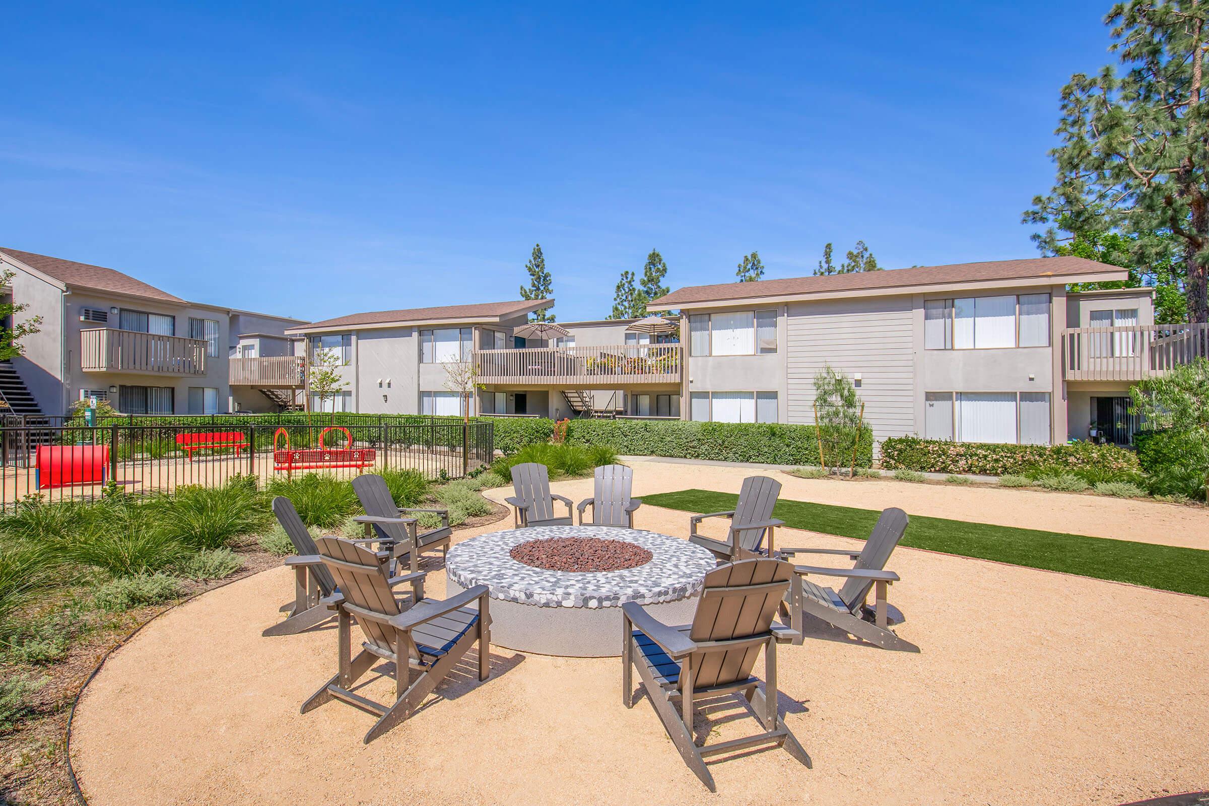 A cozy outdoor area featuring a circular fire pit surrounded by wooden Adirondack chairs, with landscaped greenery and colorful play equipment in the background. The buildings have balconies and are set against a clear blue sky, creating a welcoming community atmosphere.