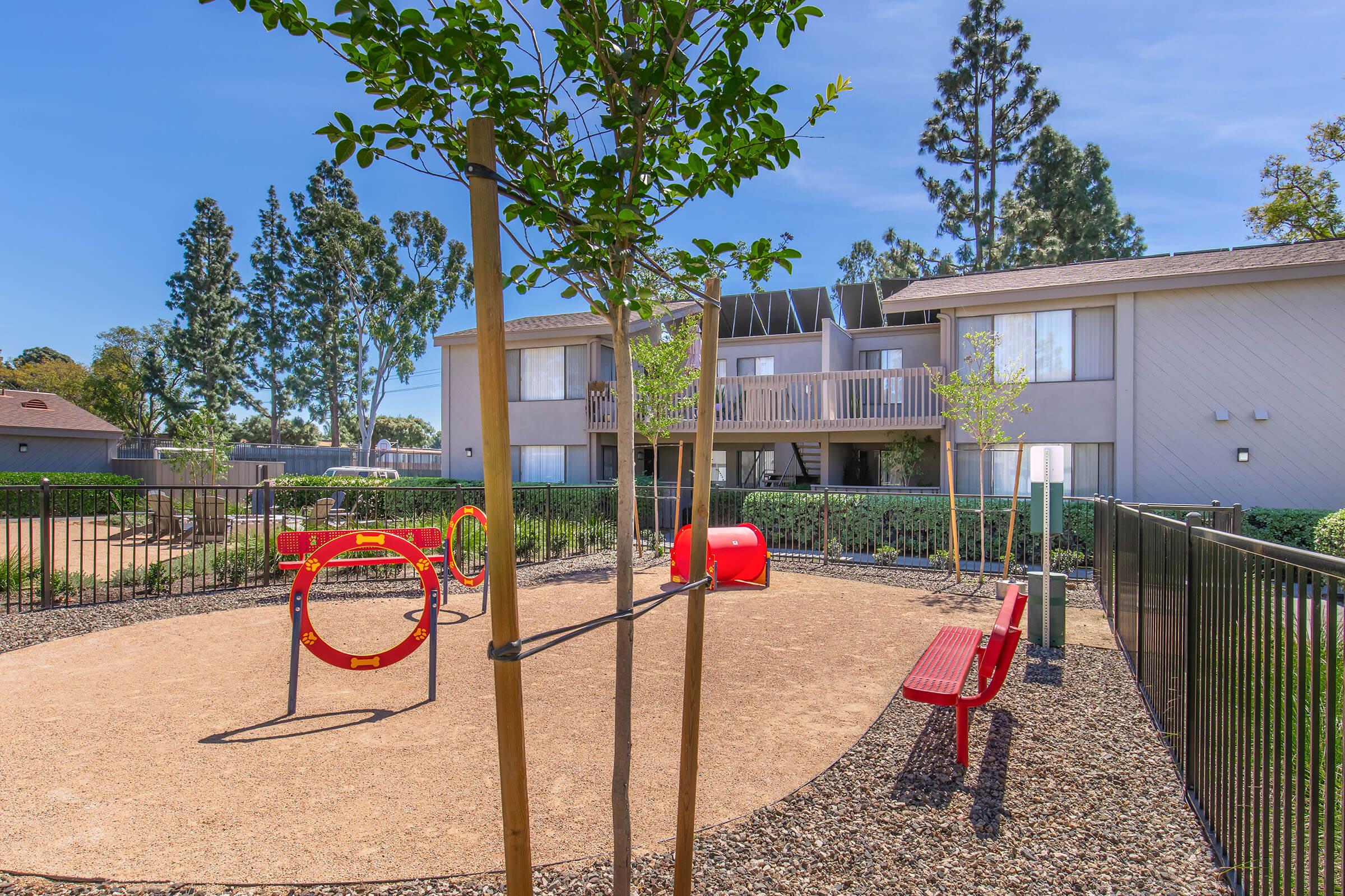 A small playground area featuring a red swing set, circular climbing structure, and benches, surrounded by mulch and grass. In the background, there are several trees and a residential building with balconies. The scene is bright and sunny, suggesting a cheerful outdoor space for children.