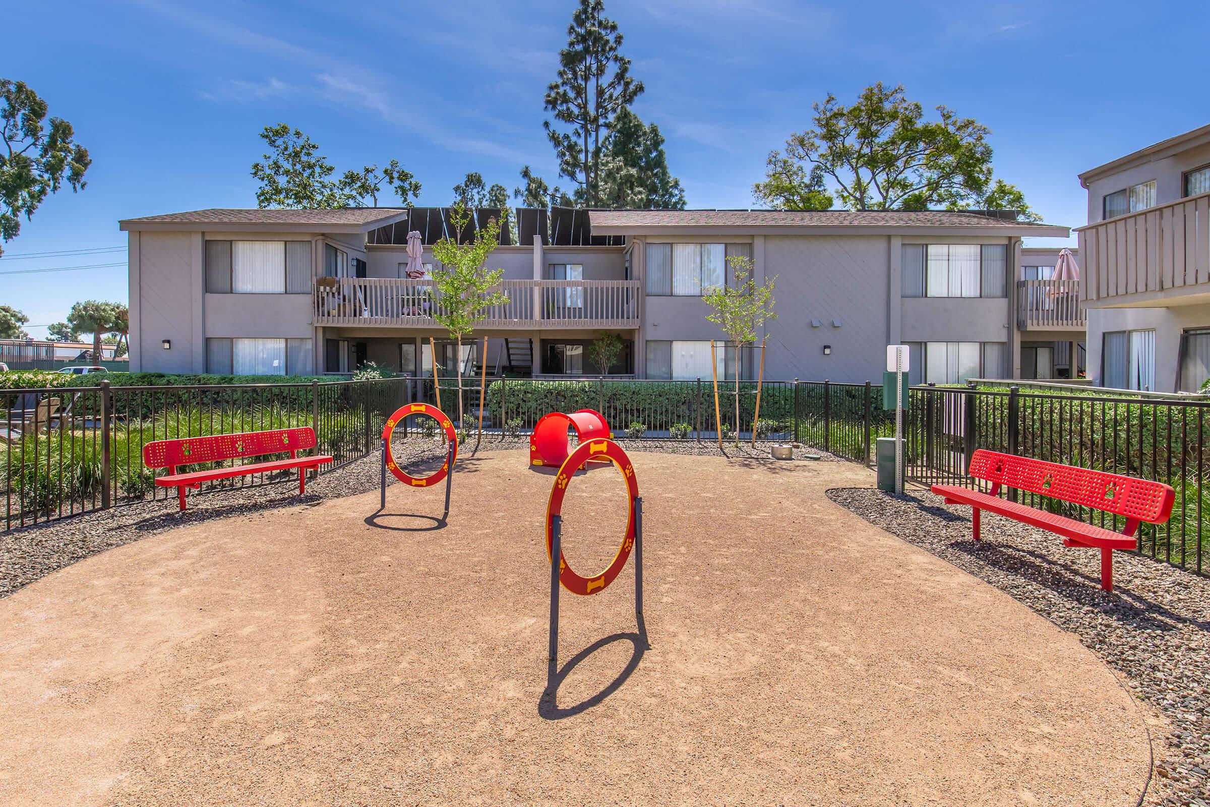 A playground area featuring two red circular play structures, surrounded by gravel and bordered by a black fence. There are two red benches on either side, and in the background, two multi-story apartment buildings are visible, with greenery and trees nearby under a clear blue sky.