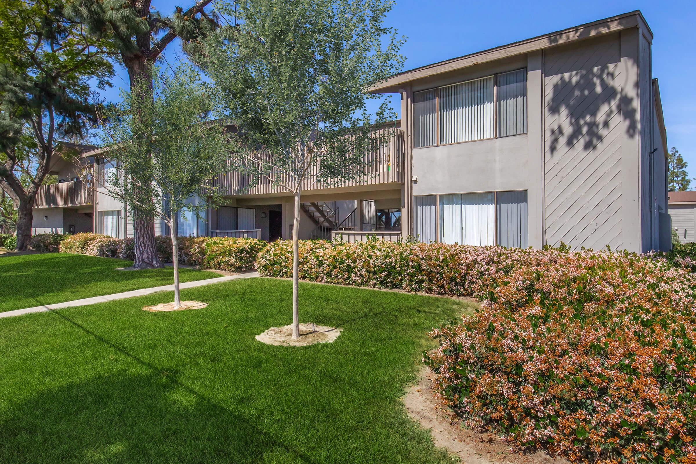 Two-story apartment building with a light gray exterior, surrounded by neatly manicured lawns and colorful flowering bushes. Small trees are planted in the grass, adding greenery to the landscape. Bright sunlight highlights the scene, creating a welcoming atmosphere.