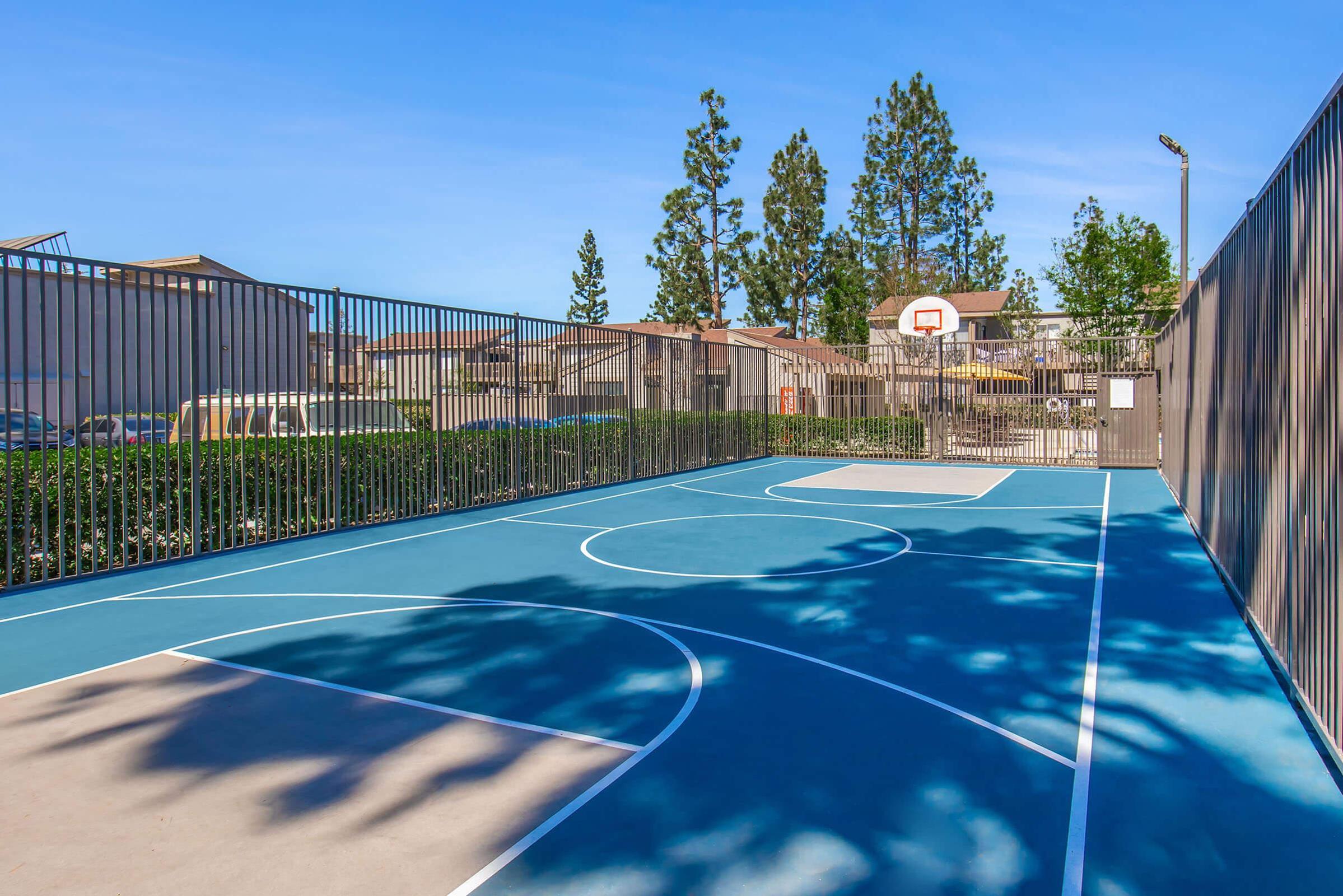 A brightly lit outdoor basketball court featuring a blue surface with white court markings. The court is enclosed by a fence, surrounded by green shrubs and trees in the background, with a basketball hoop visible at one end. Clear skies are overhead, indicating good weather.
