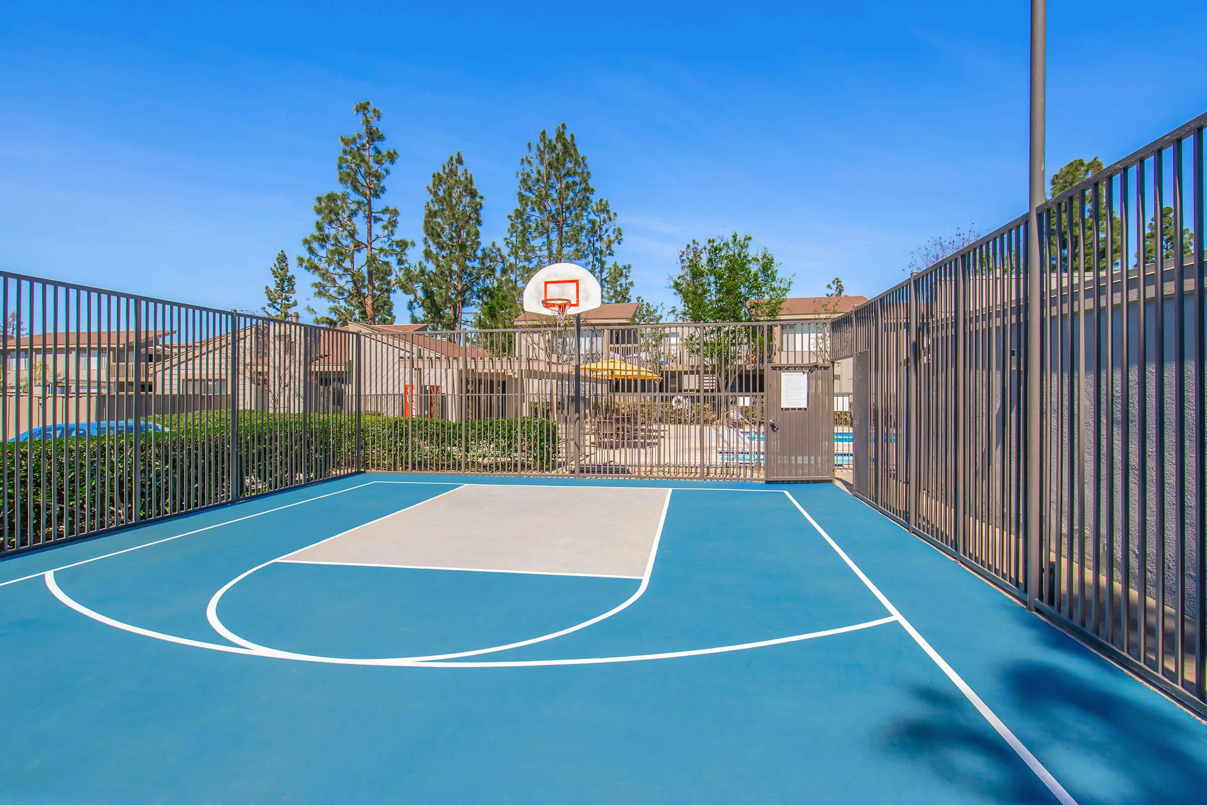 A fenced basketball court featuring a blue playing surface, a white three-point line, and a hoop. Surrounding the court are trees and buildings, with a clear blue sky overhead. The setting suggests a recreational area or community space.
