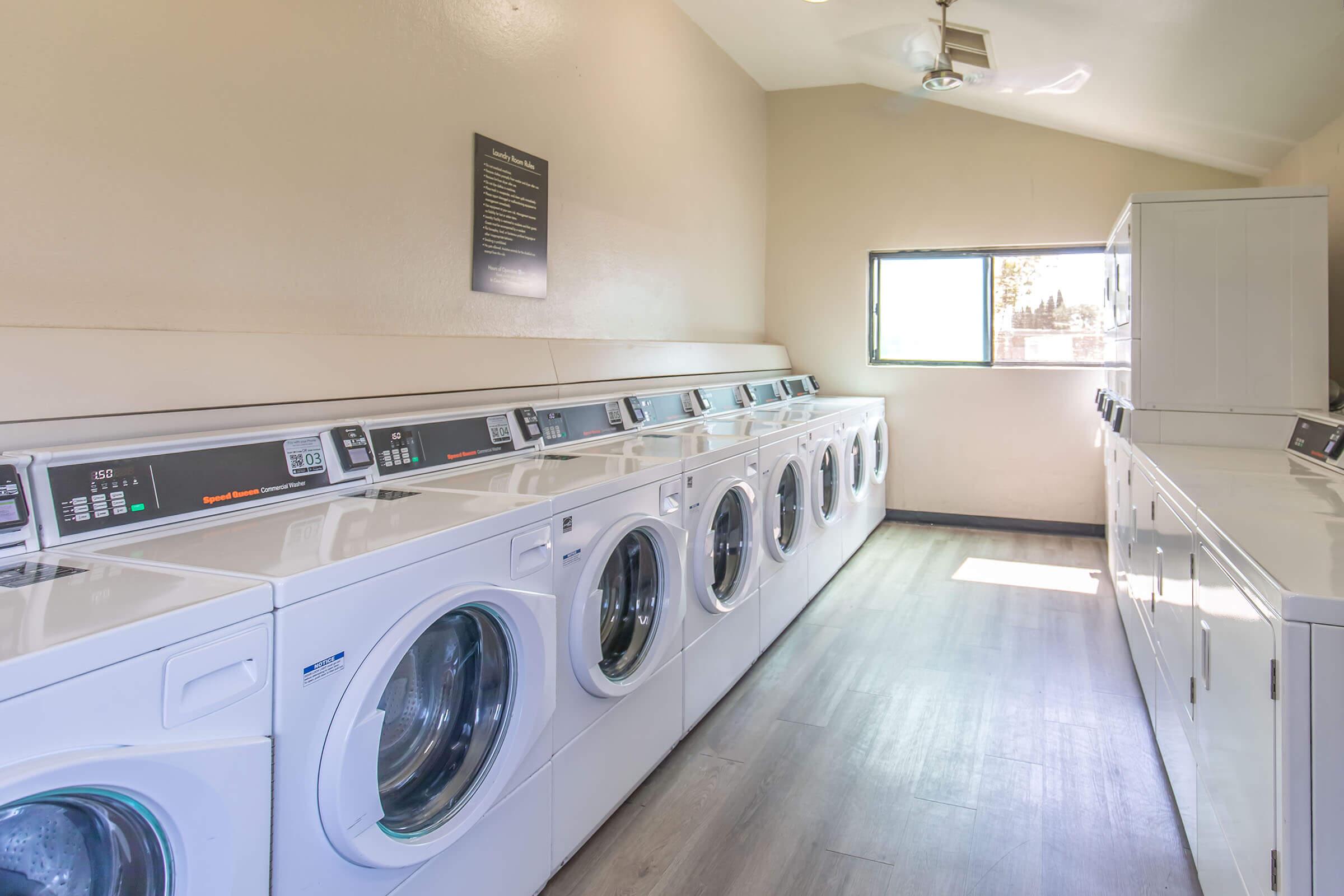 A bright and clean laundry room featuring a row of stacked washing machines and dryers along the wall. The space has light-colored walls, a window allowing natural light, and a ceiling fan. A sign is mounted on the wall, providing information about the facilities. The flooring is light wood.