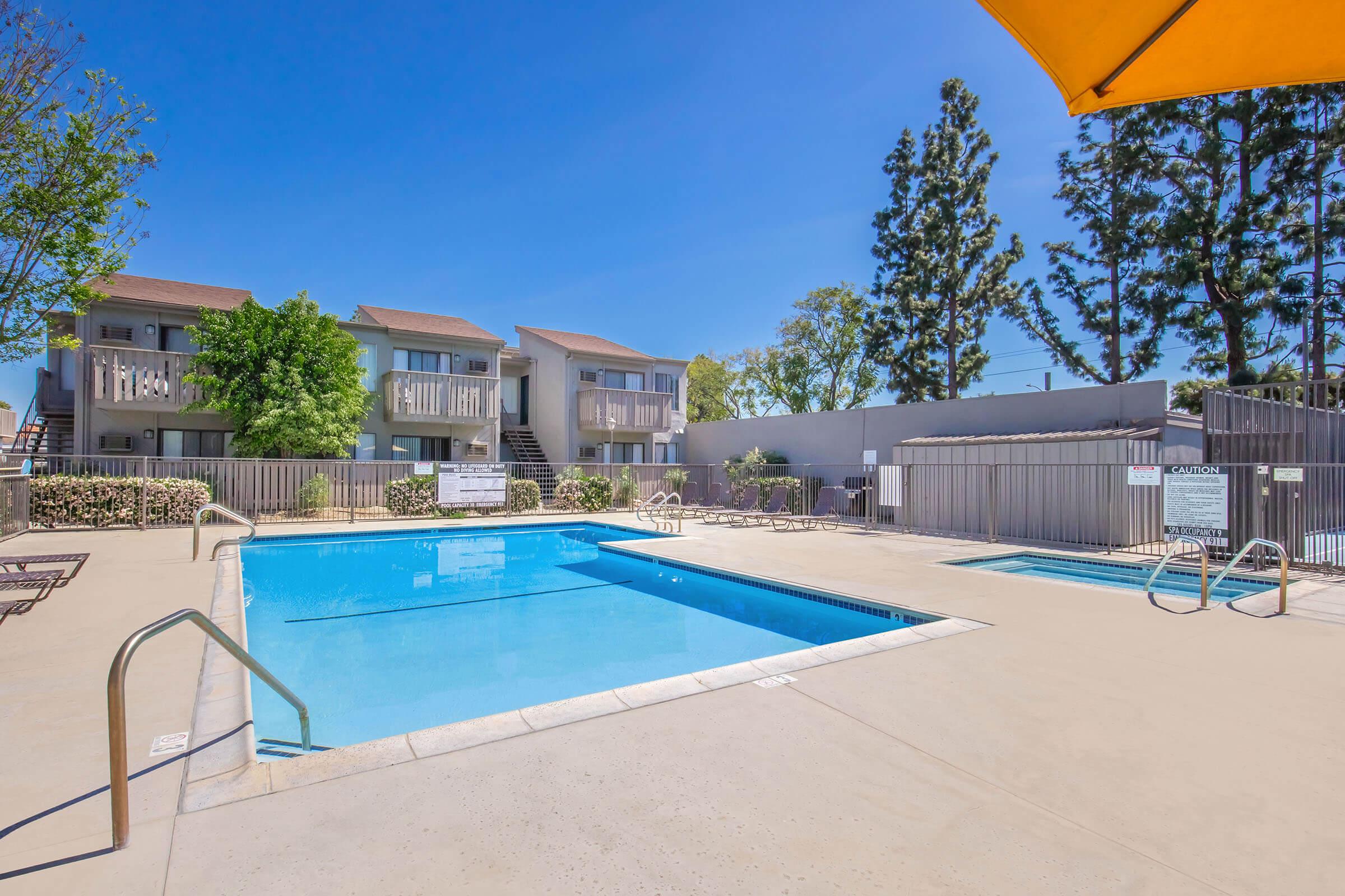 A clear blue swimming pool surrounded by a patio area, with lounge chairs and umbrellas. In the background, there are several two-story apartment buildings and trees providing shade. The sky is bright and sunny, indicating a warm day.