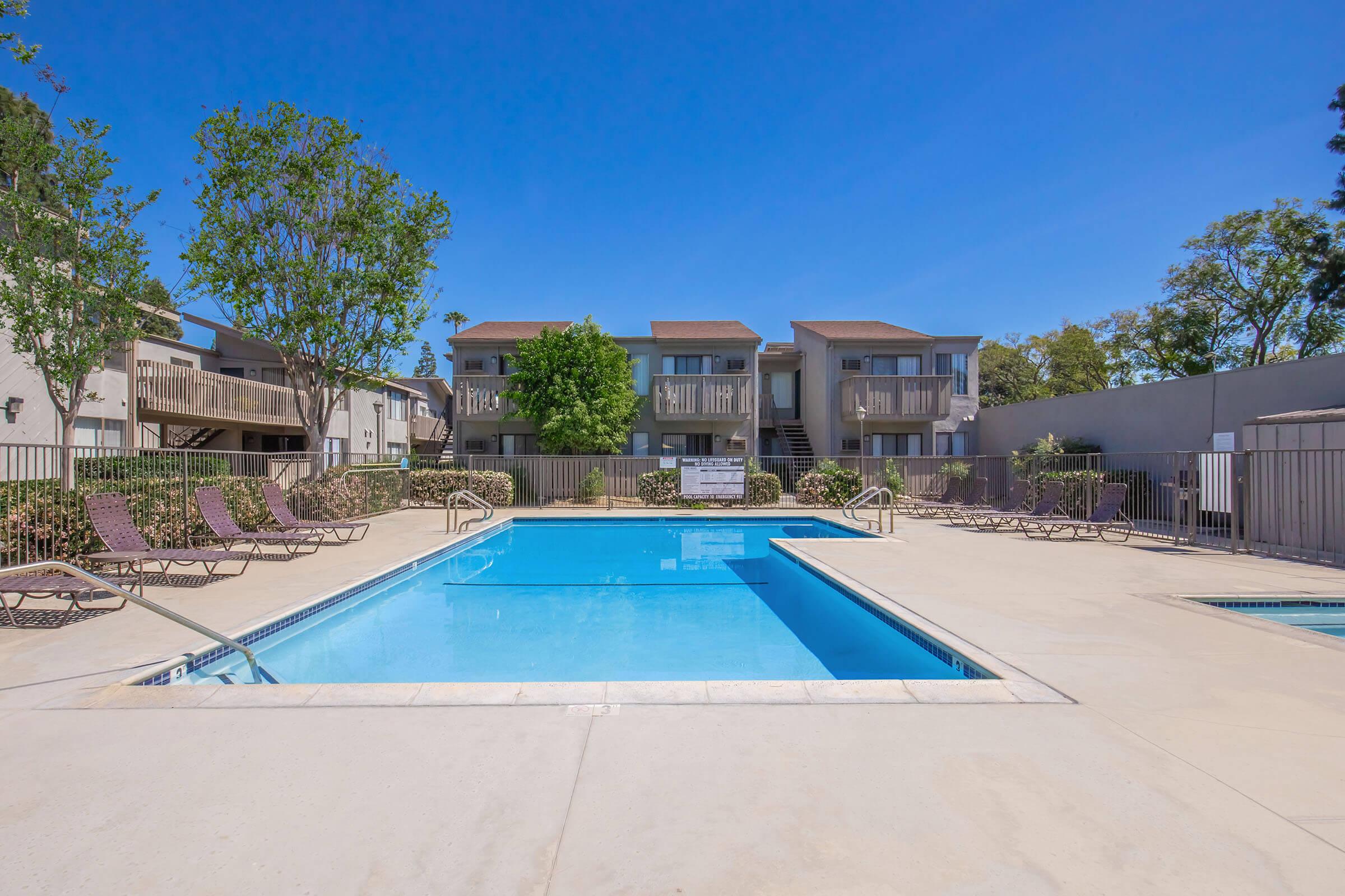 A clean, bright swimming pool surrounded by lounge chairs, set in a sunny courtyard with trees and apartment buildings in the background. The scene features clear blue skies and a well-maintained area, ideal for relaxation and leisure.