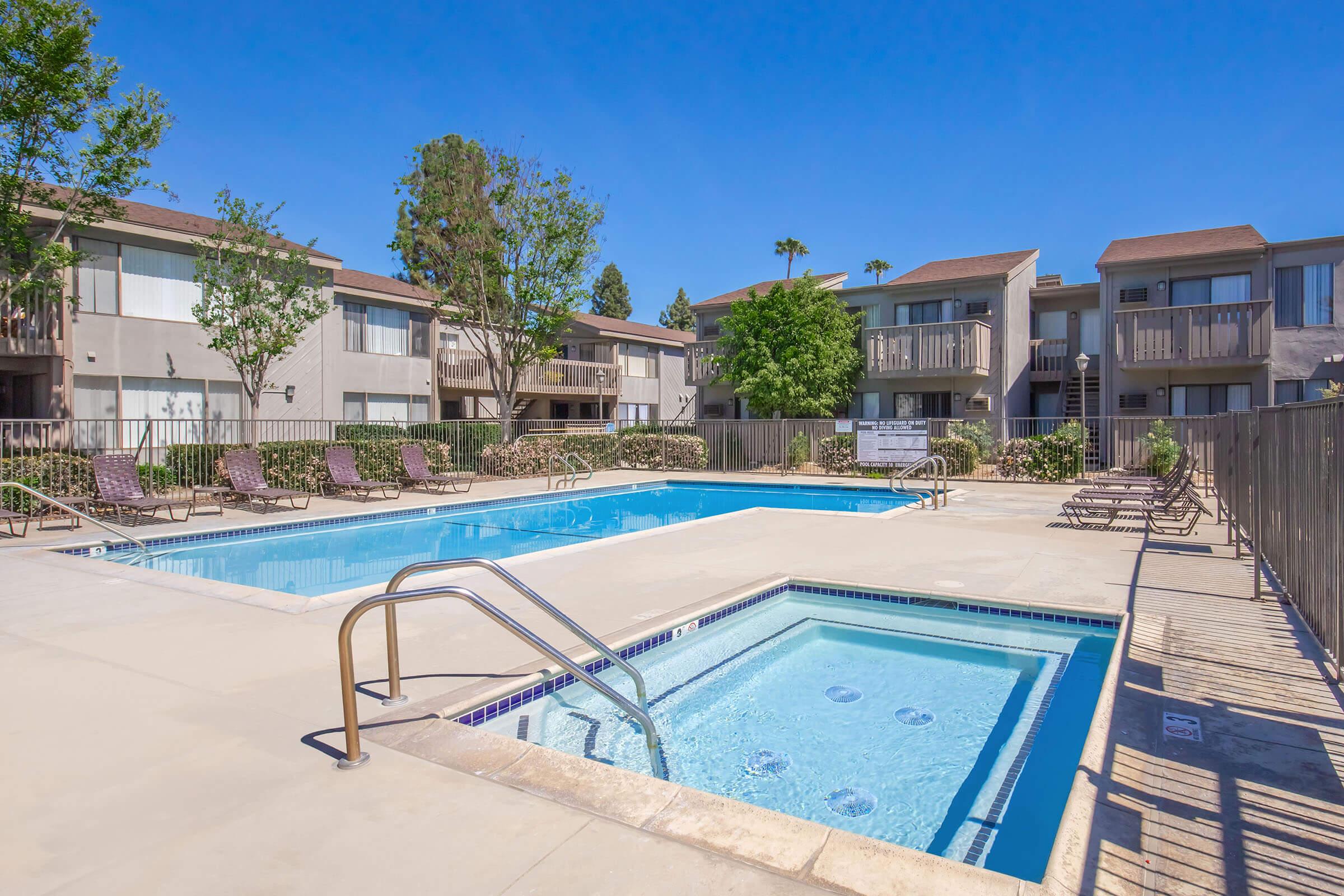 A sunny outdoor pool area featuring a large swimming pool and a separate hot tub. Surrounding the pool are lounge chairs, with landscaped greenery and apartment buildings in the background under a clear blue sky.